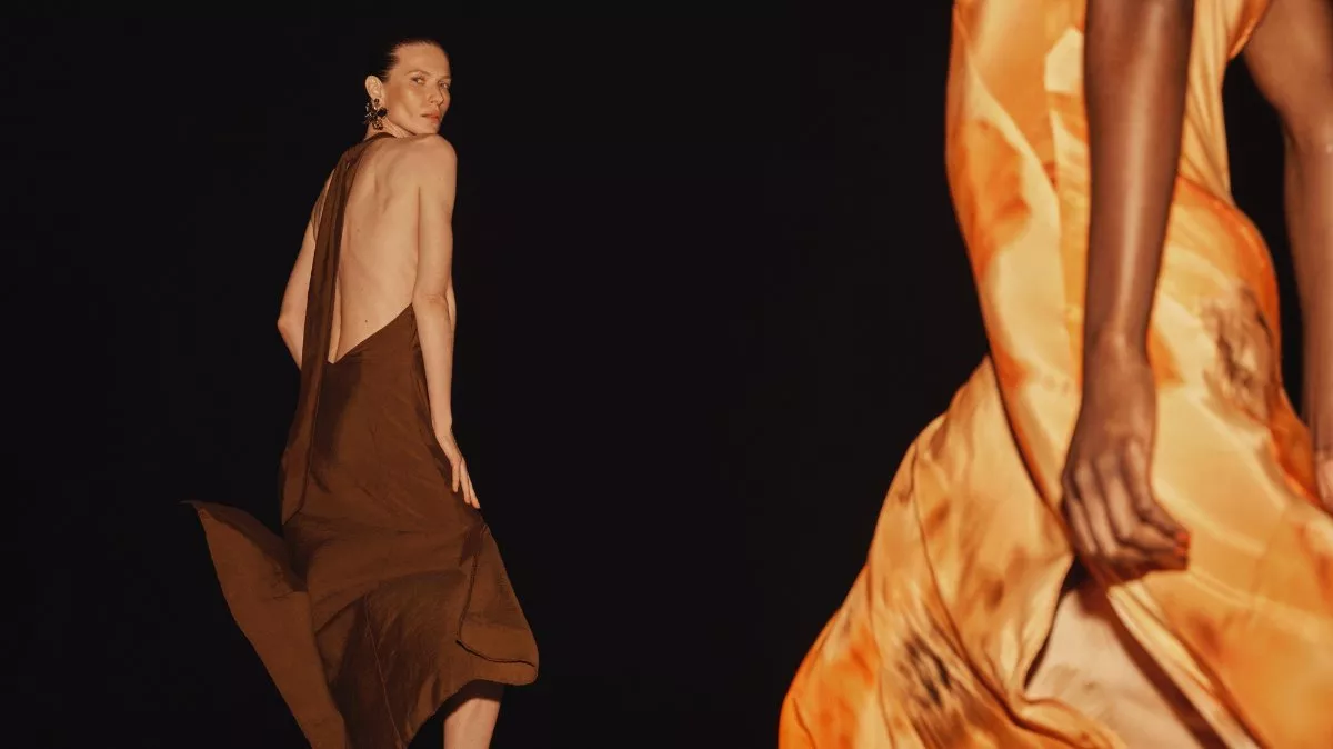 Fashion runway model in backless brown dress, with blurred orange gown foreground on black backdrop