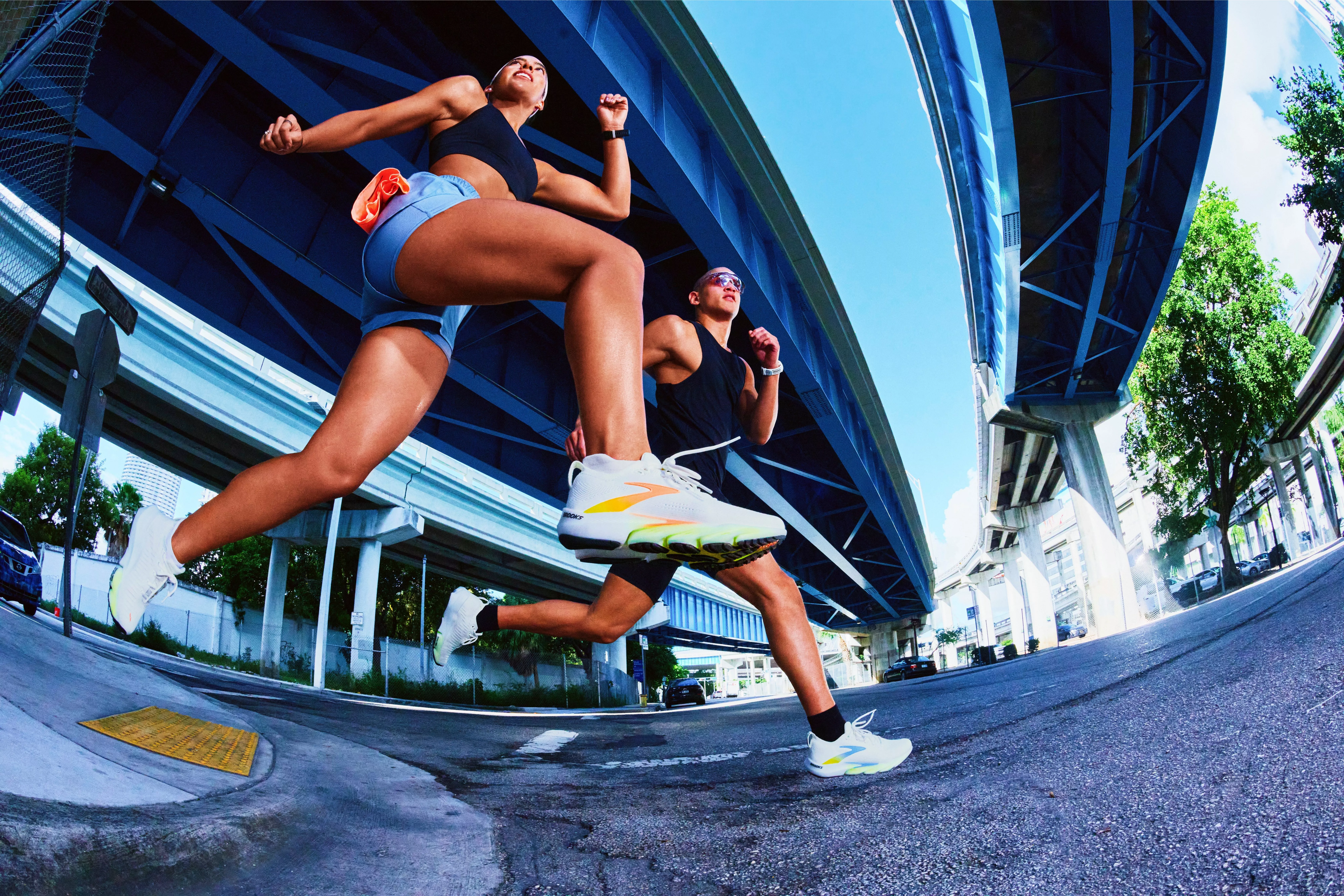 Two runners sprinting under city flyovers on an urban street, low-angle action shot in bright daylight