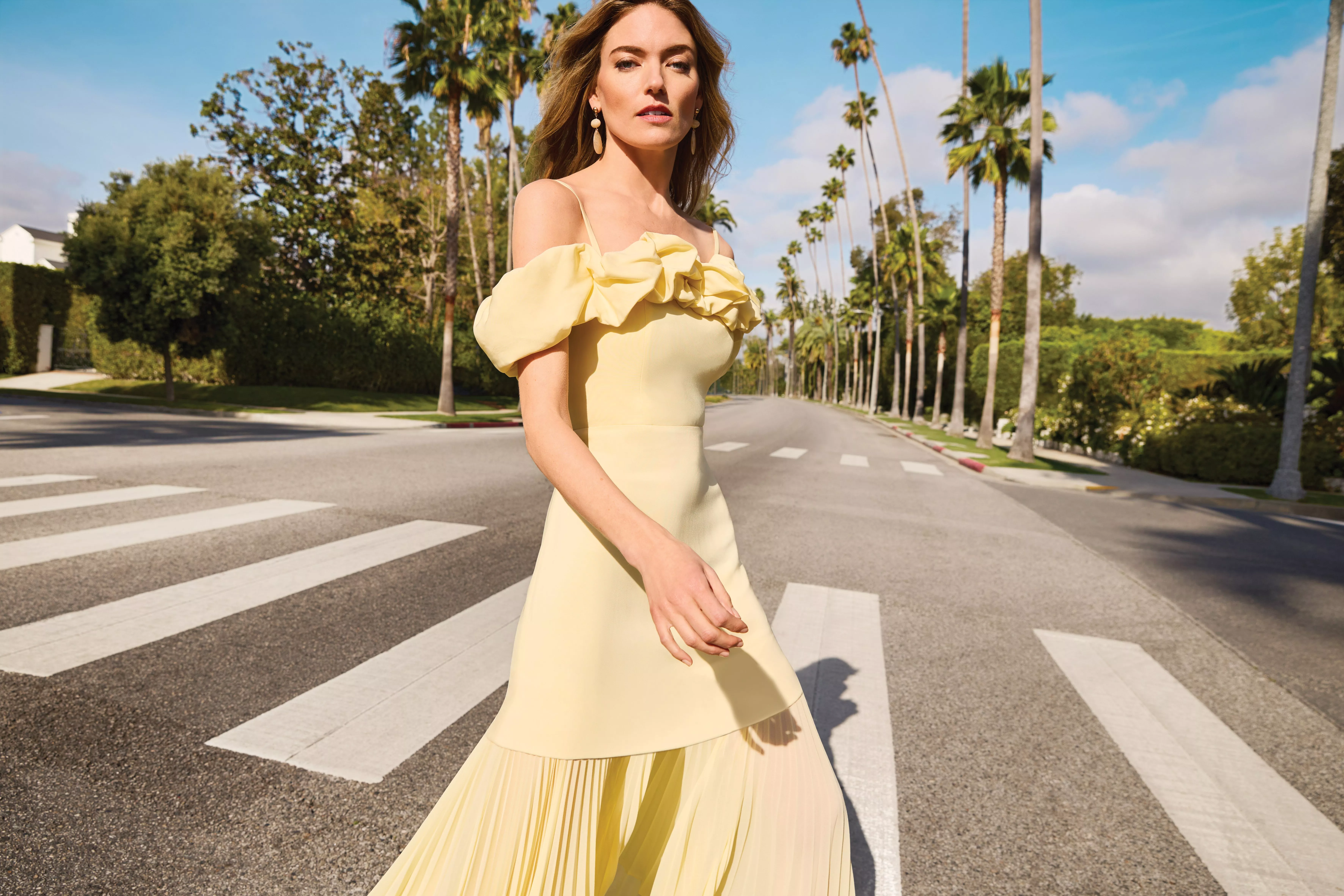 Woman in pale yellow off-shoulder dress walking on palm-lined street, sunny luxury fashion shoot outdoors