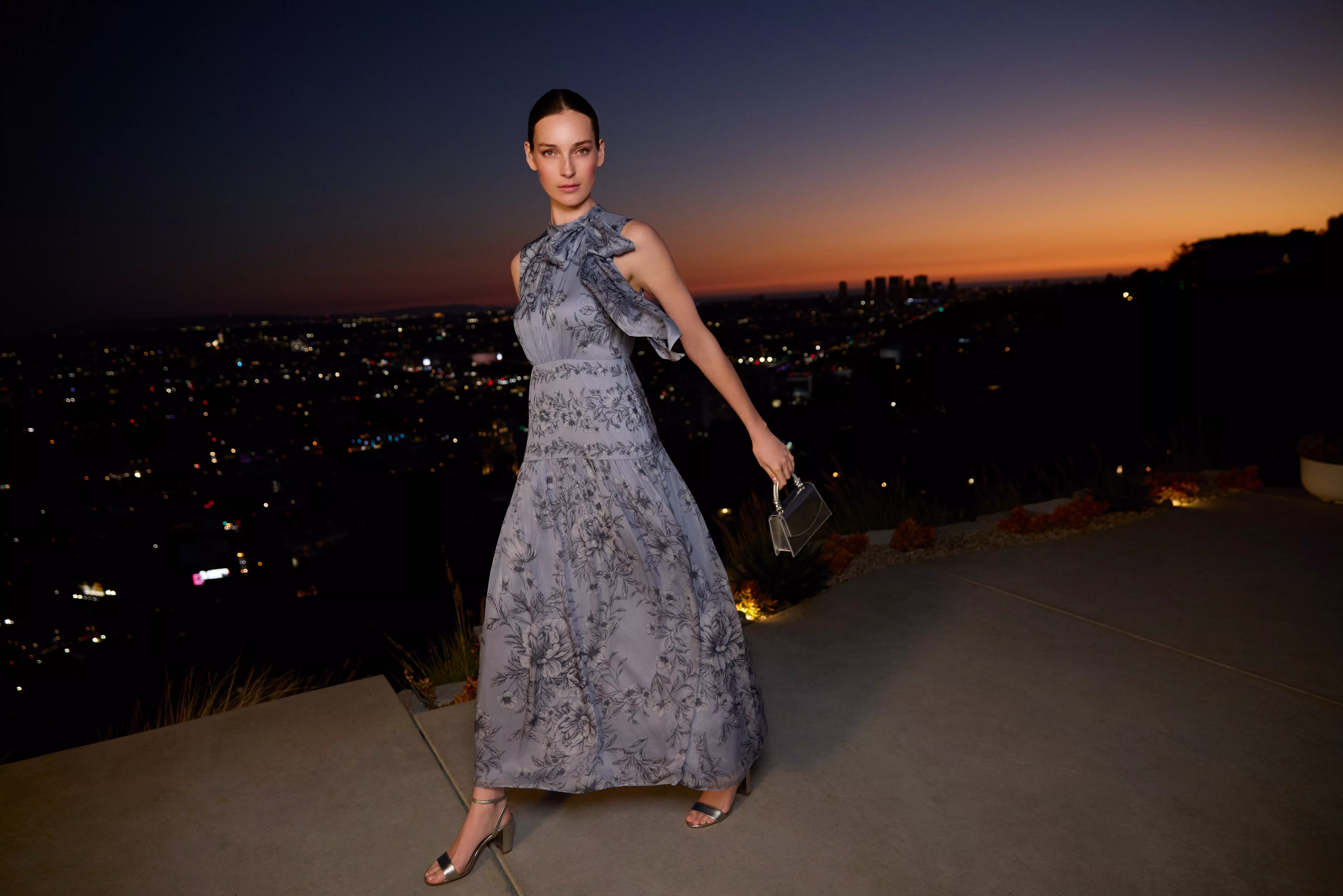 Elegant woman in grey floral evening gown holding clutch, posing at sunset with city skyline lights behind