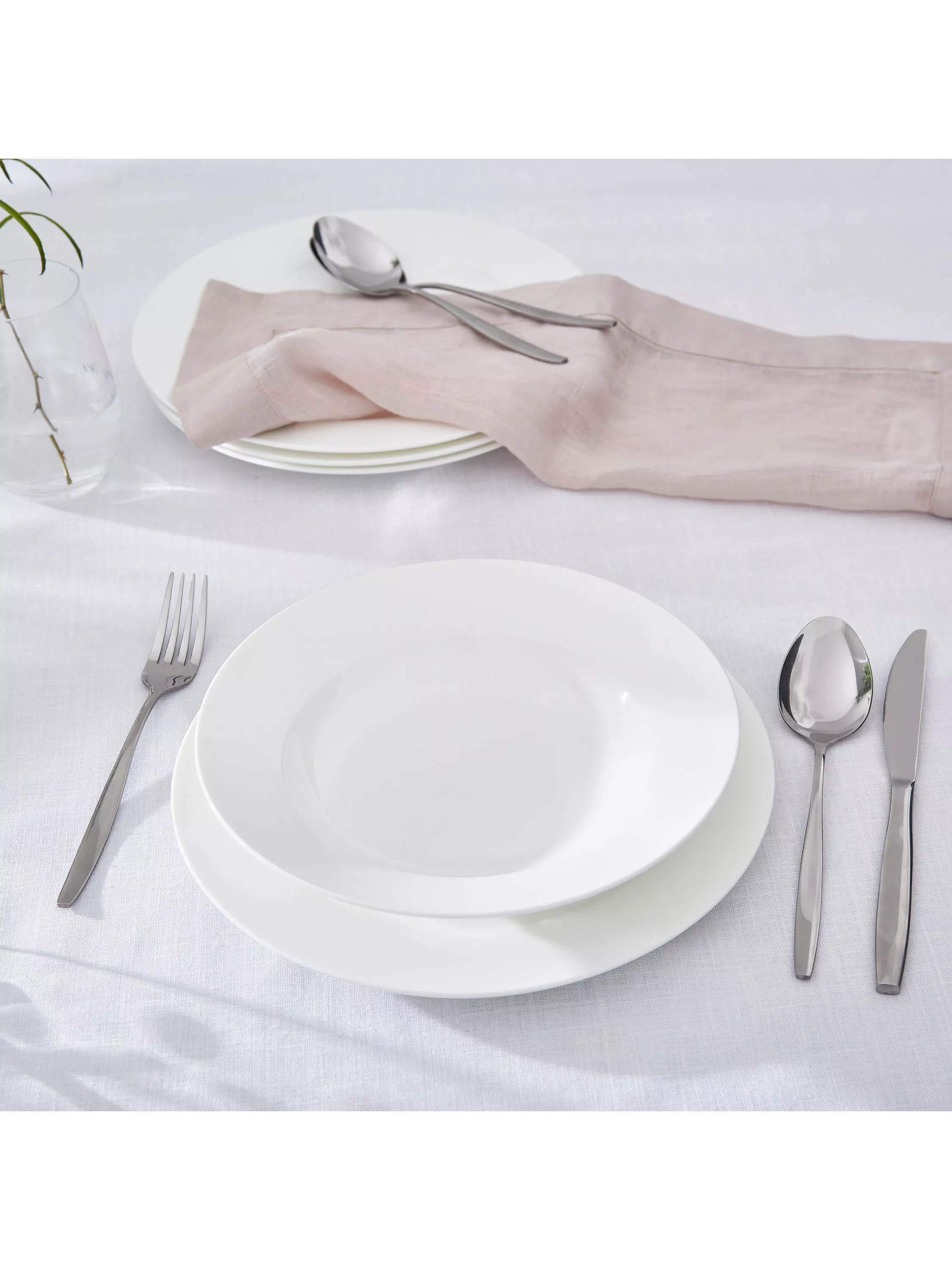 Minimal white table setting with plates, silver cutlery and blush linen napkin on crisp tablecloth for dining decor