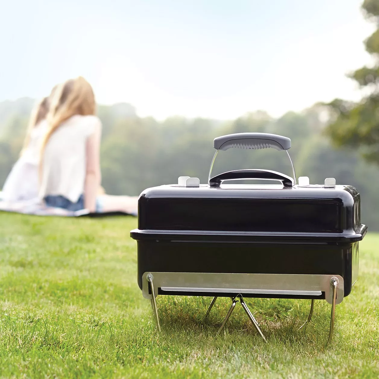 Portable charcoal barbecue grill on grass, with people relaxing in the background on a sunny day outdoors