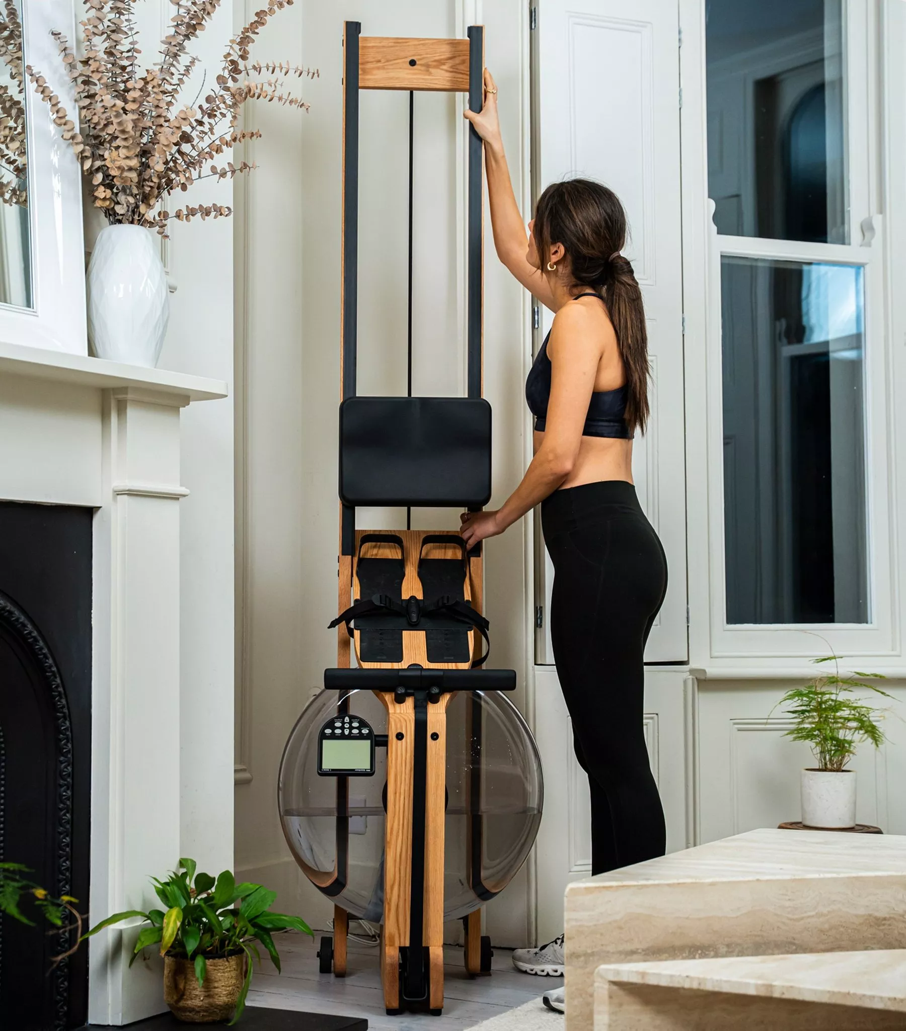 Woman folding wooden water rowing machine upright in living room, compact home gym fitness equipment storage