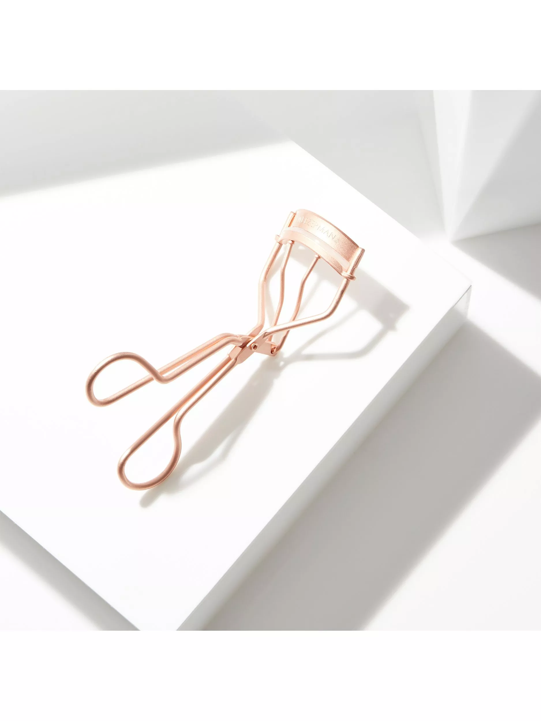 Rose gold eyelash curler on white display block with bright natural light and soft shadows, beauty tool flat lay