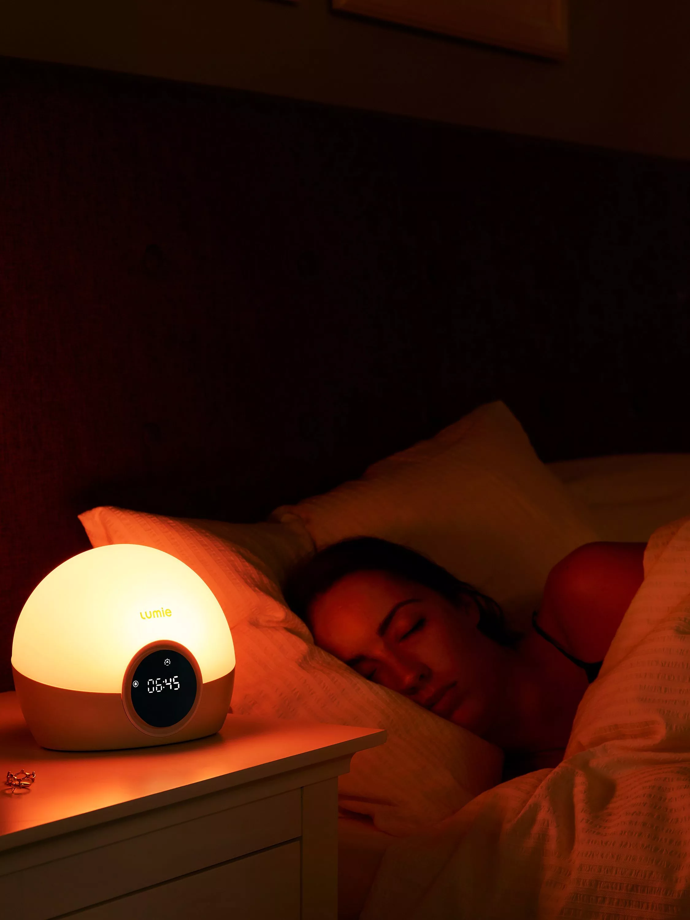 Warm bedside sunrise alarm clock lamp on nightstand beside sleeping woman in cosy bedroom at night