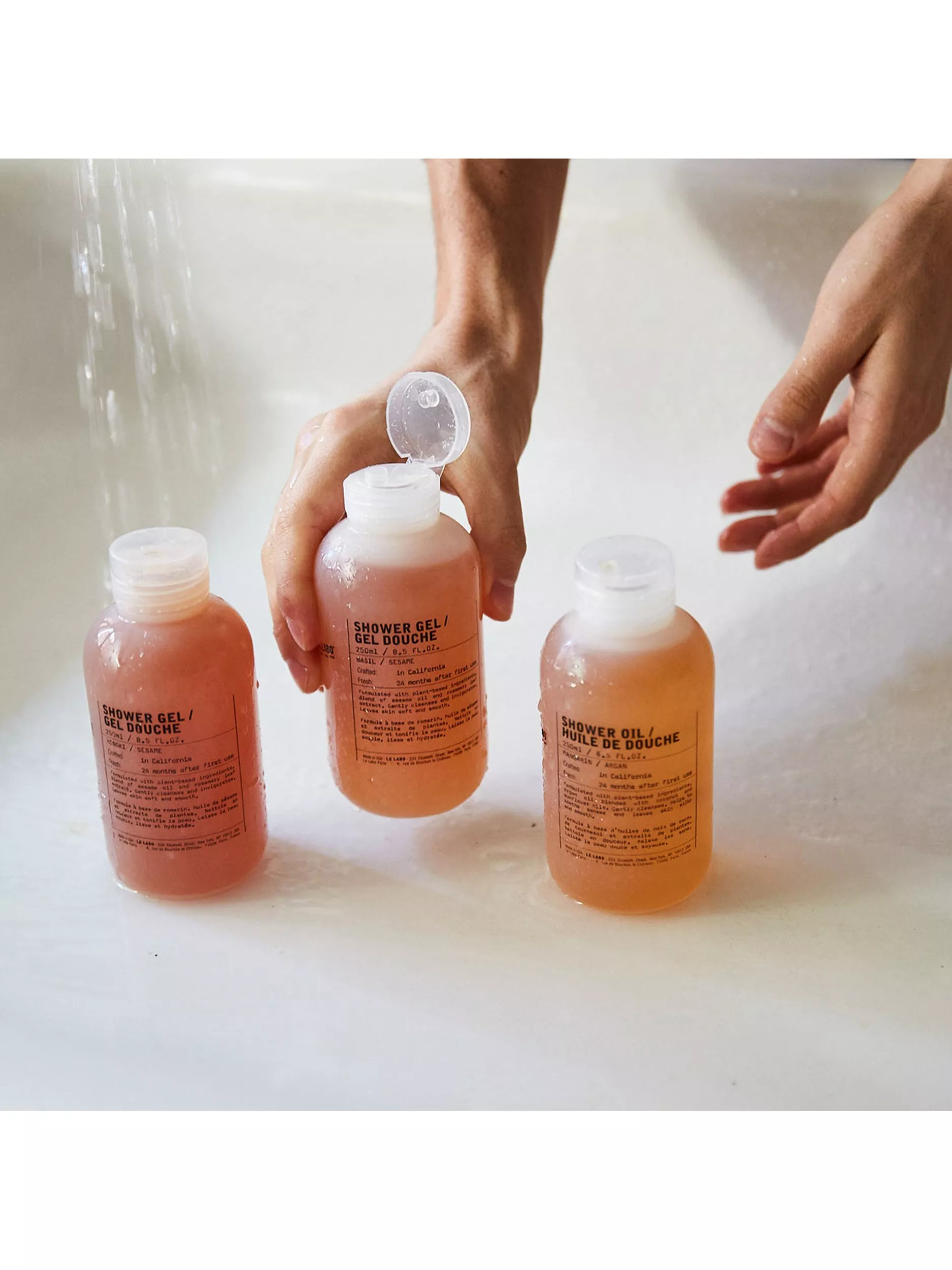 Hands holding shower gel bottle in bath, with three travel-size shower gel and shower oil toiletry bottles.