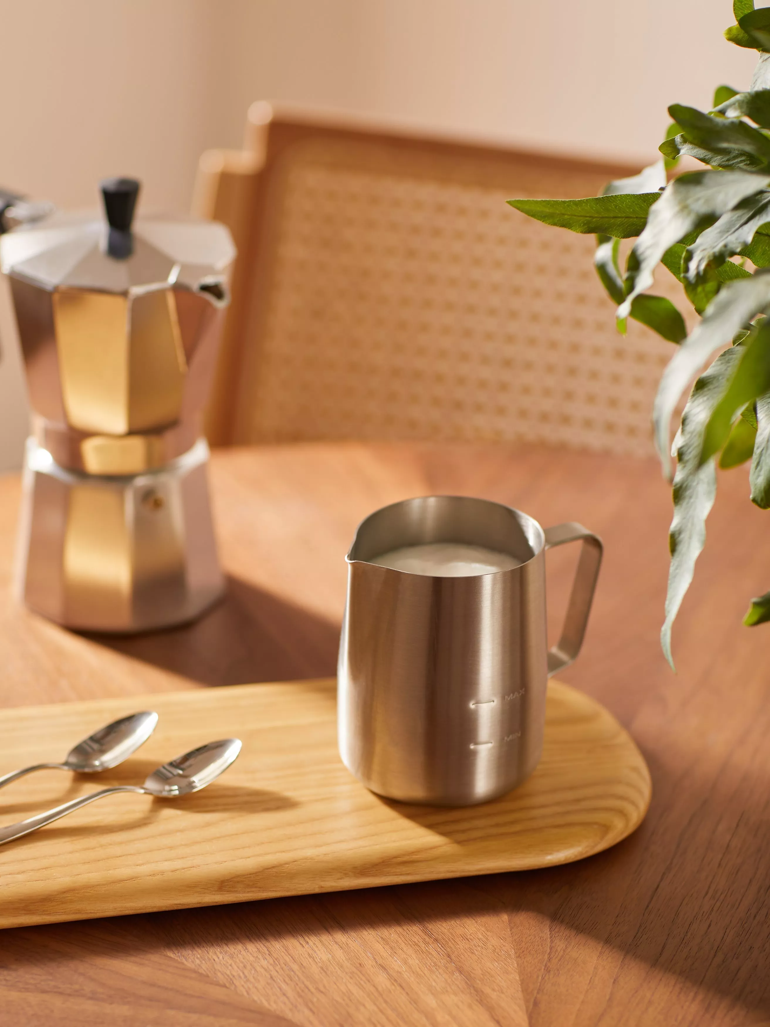 Stainless steel milk frothing jug with foam, moka pot and spoons on wooden table in cosy kitchen light