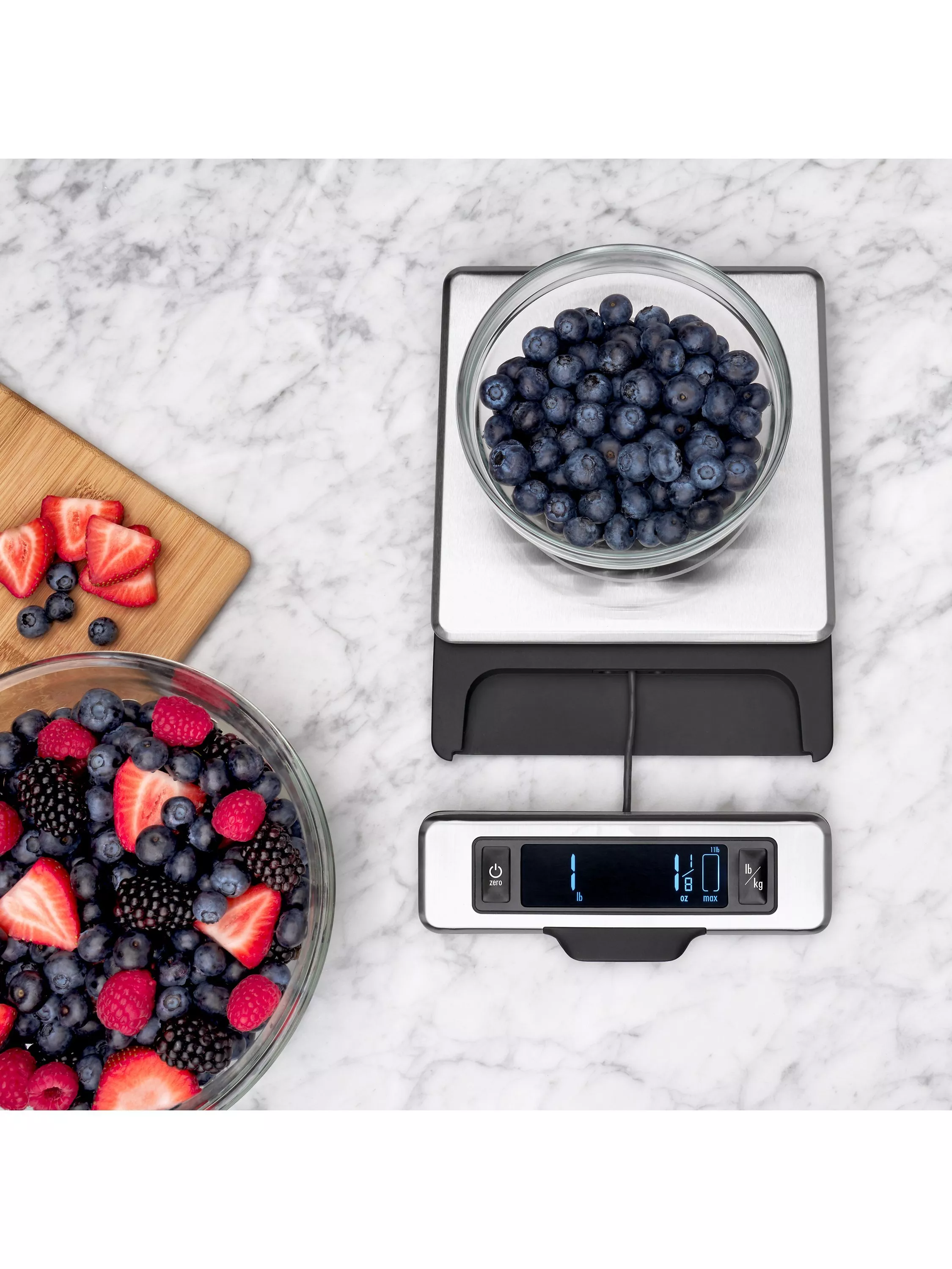 Glass bowl of fresh blueberries on digital kitchen scales, with mixed berries and sliced strawberries on marble worktop