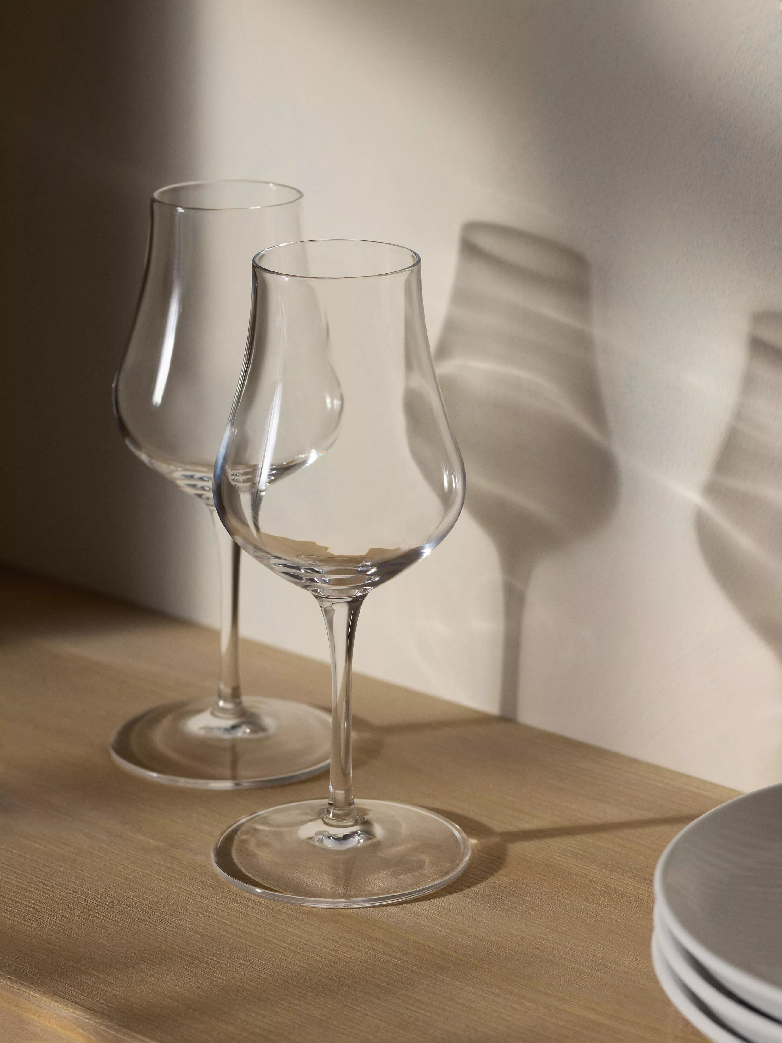 Two clear wine glasses on a wooden table, sunlit shadows on cream wall, minimal modern home décor scene