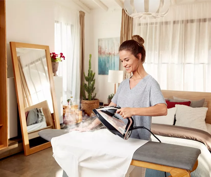 Woman steam ironing a white shirt on an ironing board in a bright modern bedroom with mirror and bed