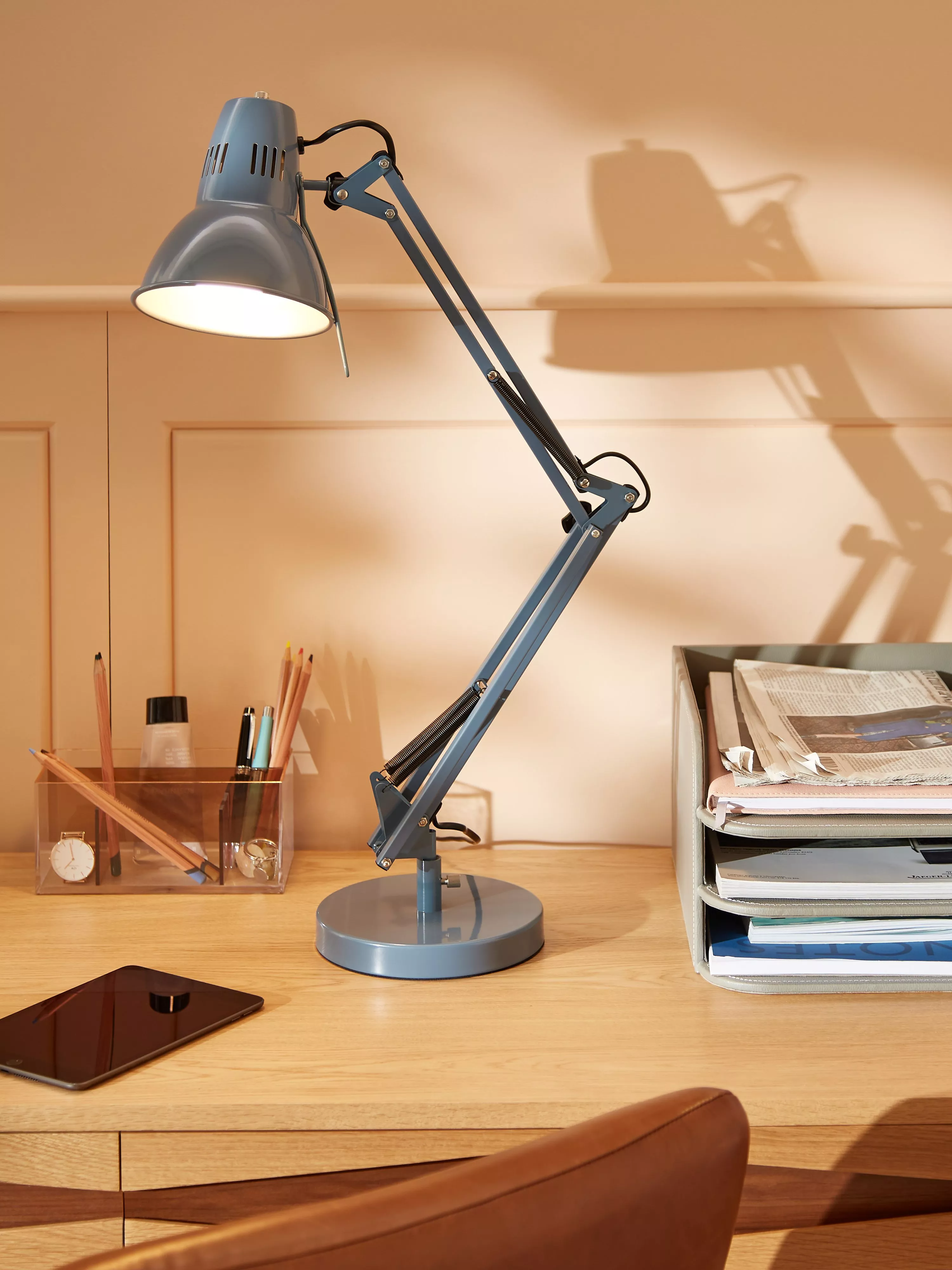 Adjustable blue desk lamp on wooden home office desk with stationery organiser, tablet and paper tray.