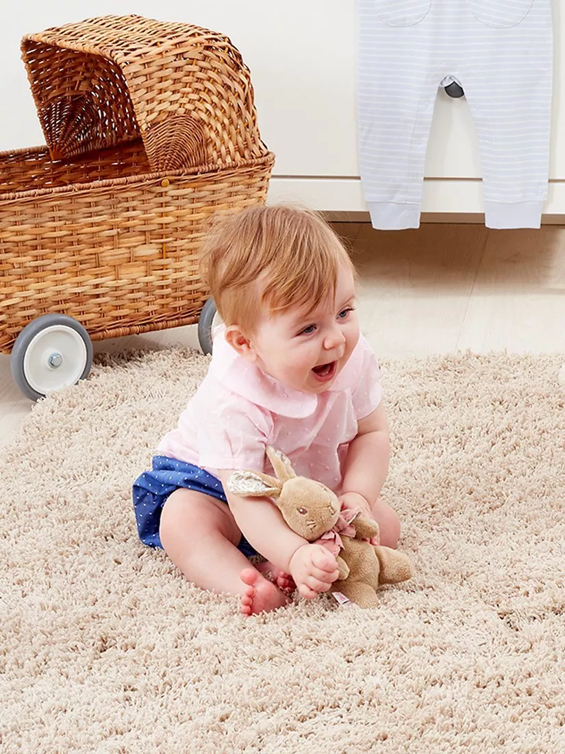 Smiling baby sitting on a soft nursery rug, cuddling a plush bunny beside a wicker toy pram indoors.