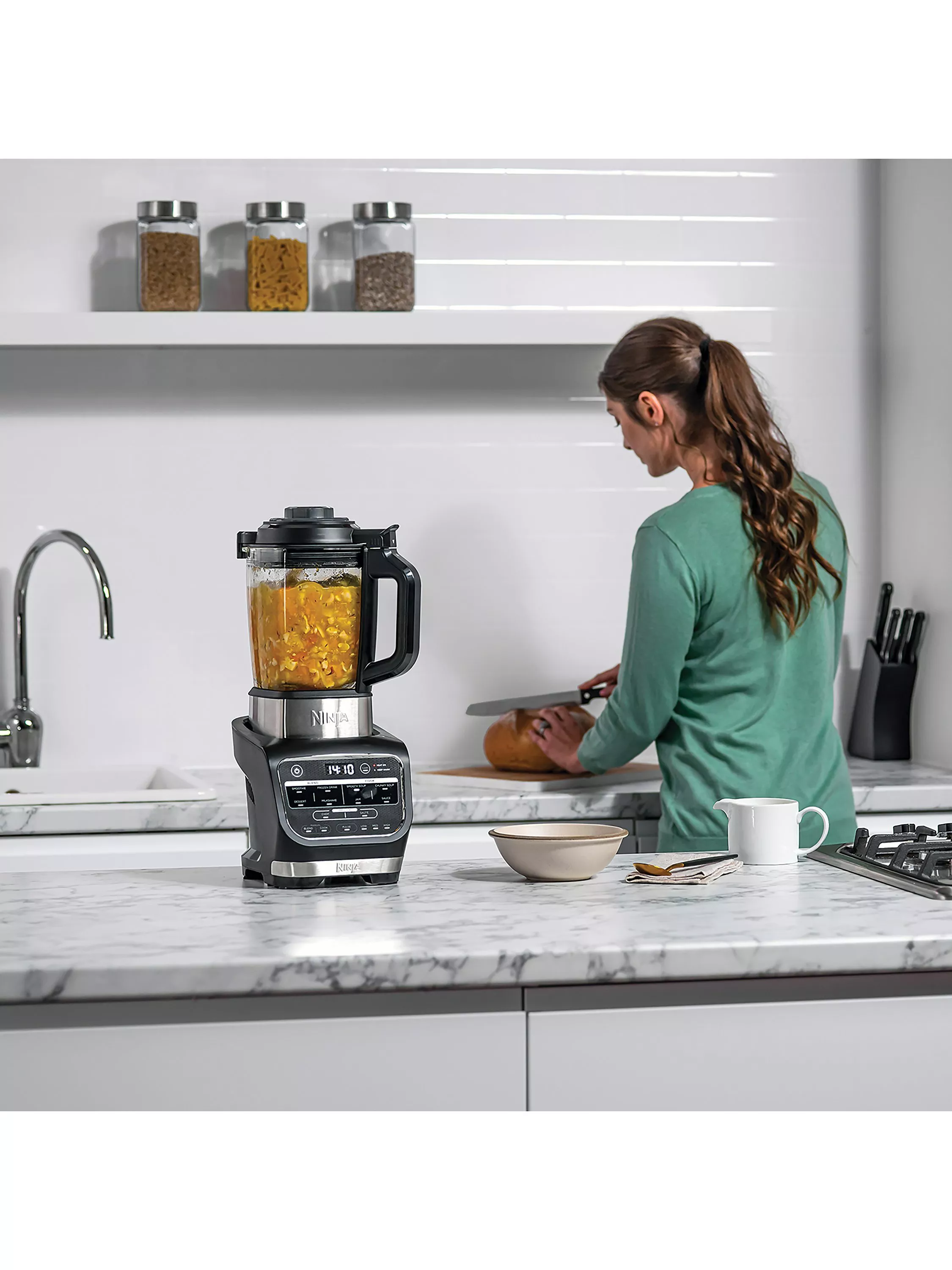 Woman preparing food in a modern kitchen beside a Ninja blender filled with soup ingredients on a marble worktop