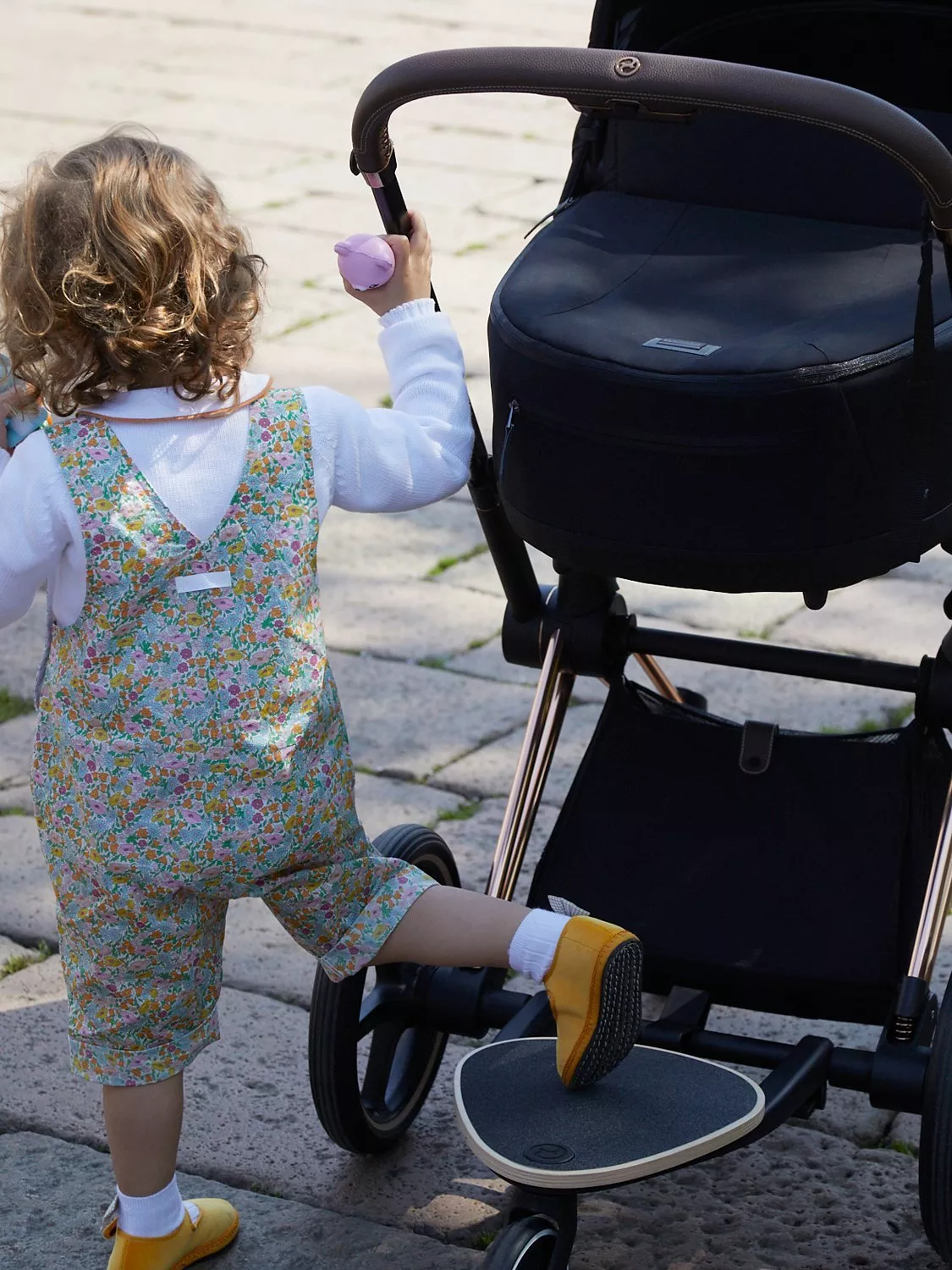 Toddler pushing a black pram with buggy board on a paved walkway, wearing floral dungarees outdoors