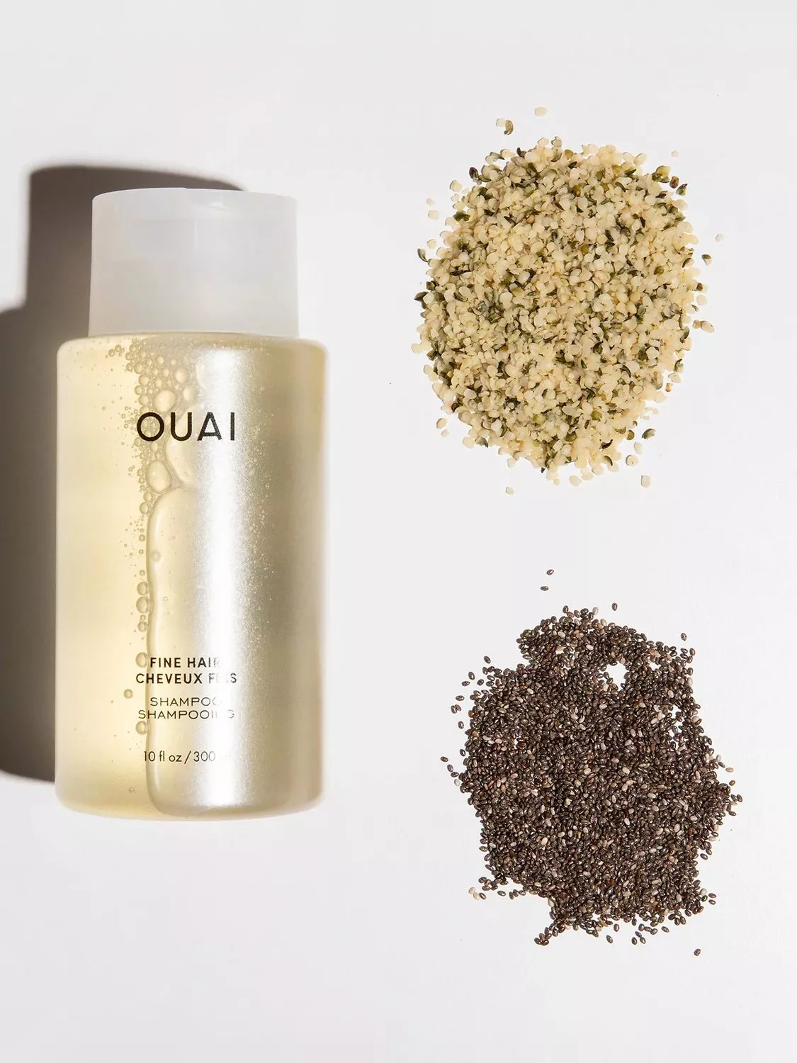 OUAI Fine Hair Shampoo bottle on white background beside hemp seeds and chia seeds, flat lay product photo
