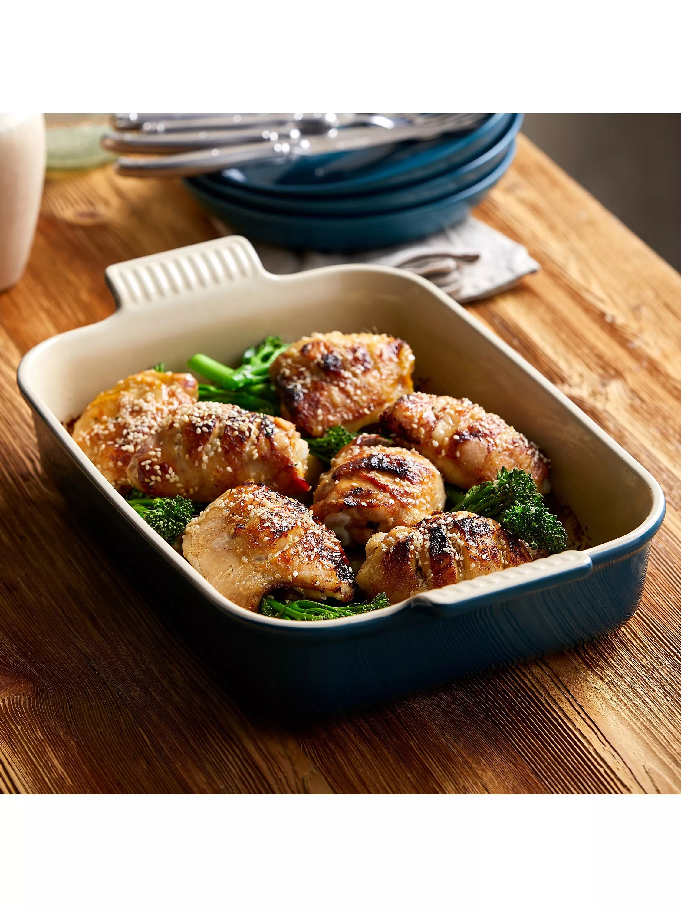 Oven-baked sesame chicken thighs with tender broccoli in a ceramic casserole dish on a rustic wooden table