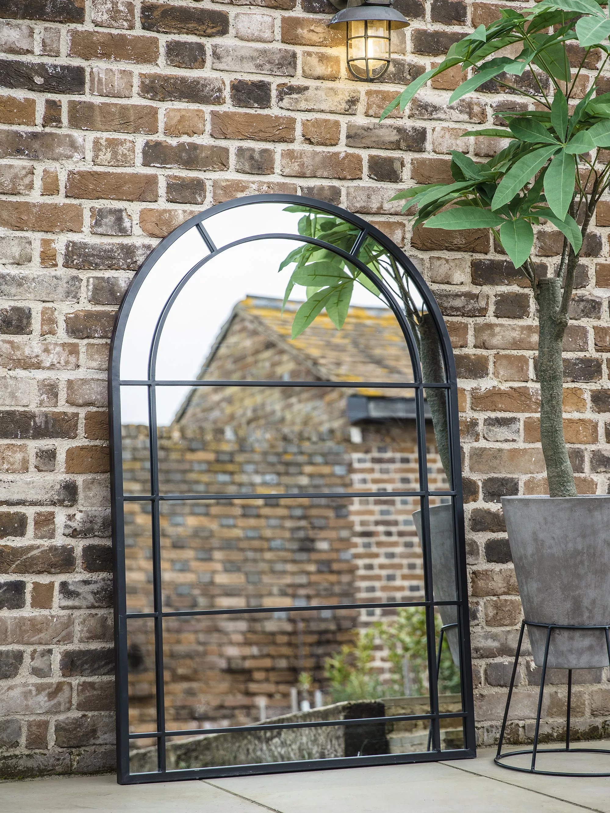 Black metal arched garden mirror against brick wall, with outdoor wall light and potted tree on patio
