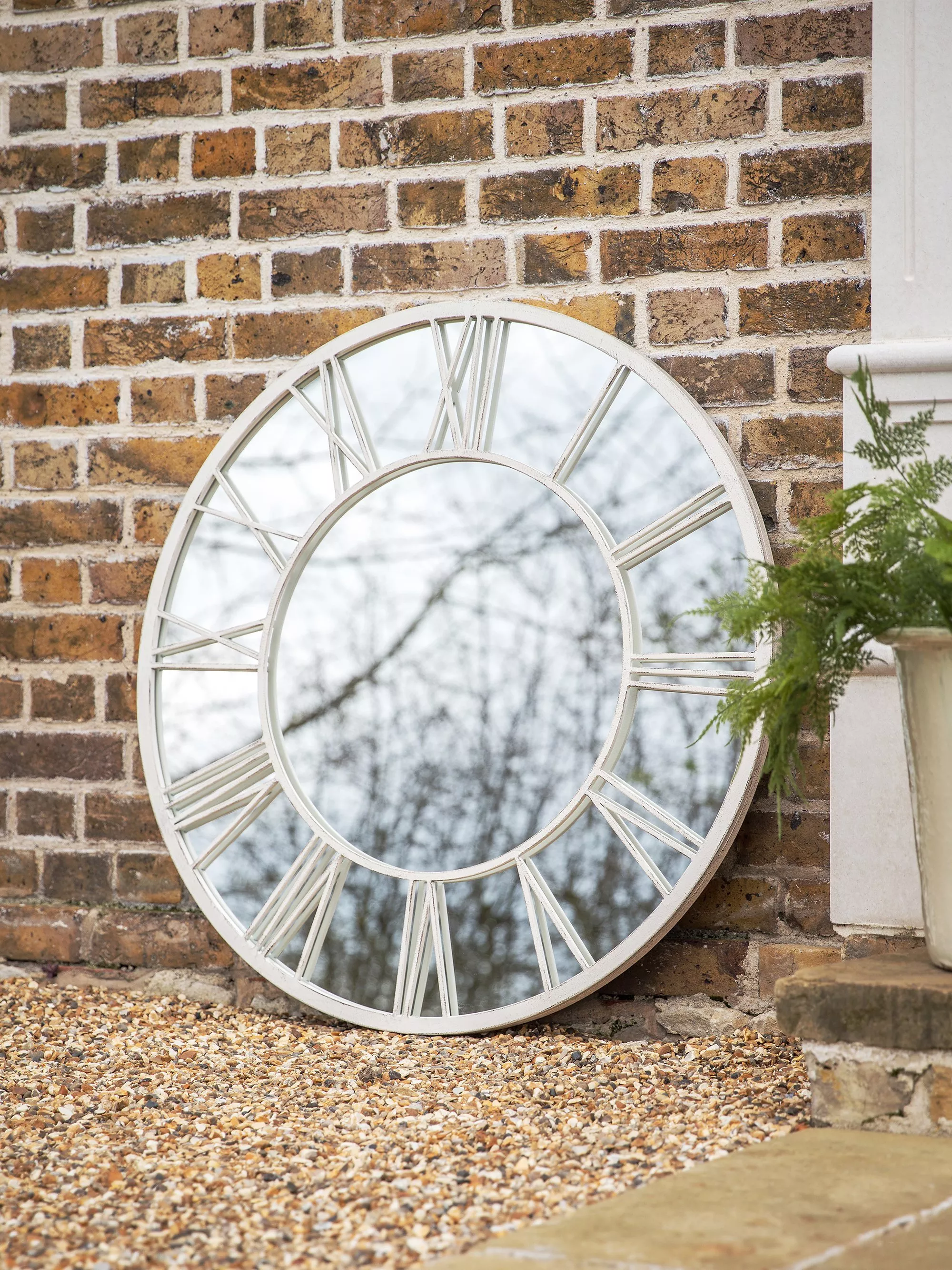 Large round white Roman numeral mirror leaning against a rustic brick wall beside a potted plant outdoors