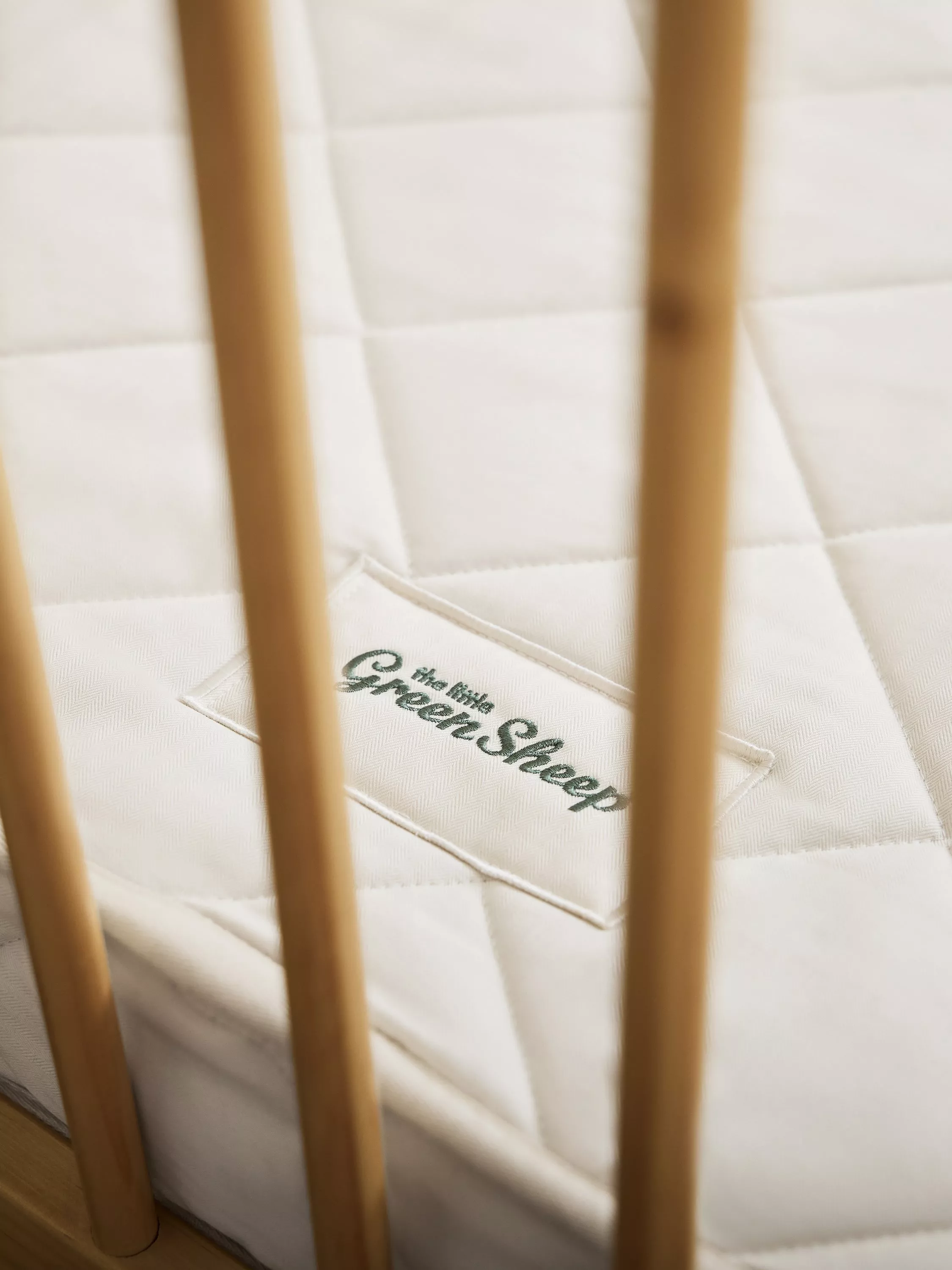 Close-up of quilted white cot mattress with GreenSheep label, seen behind natural wooden cot bars
