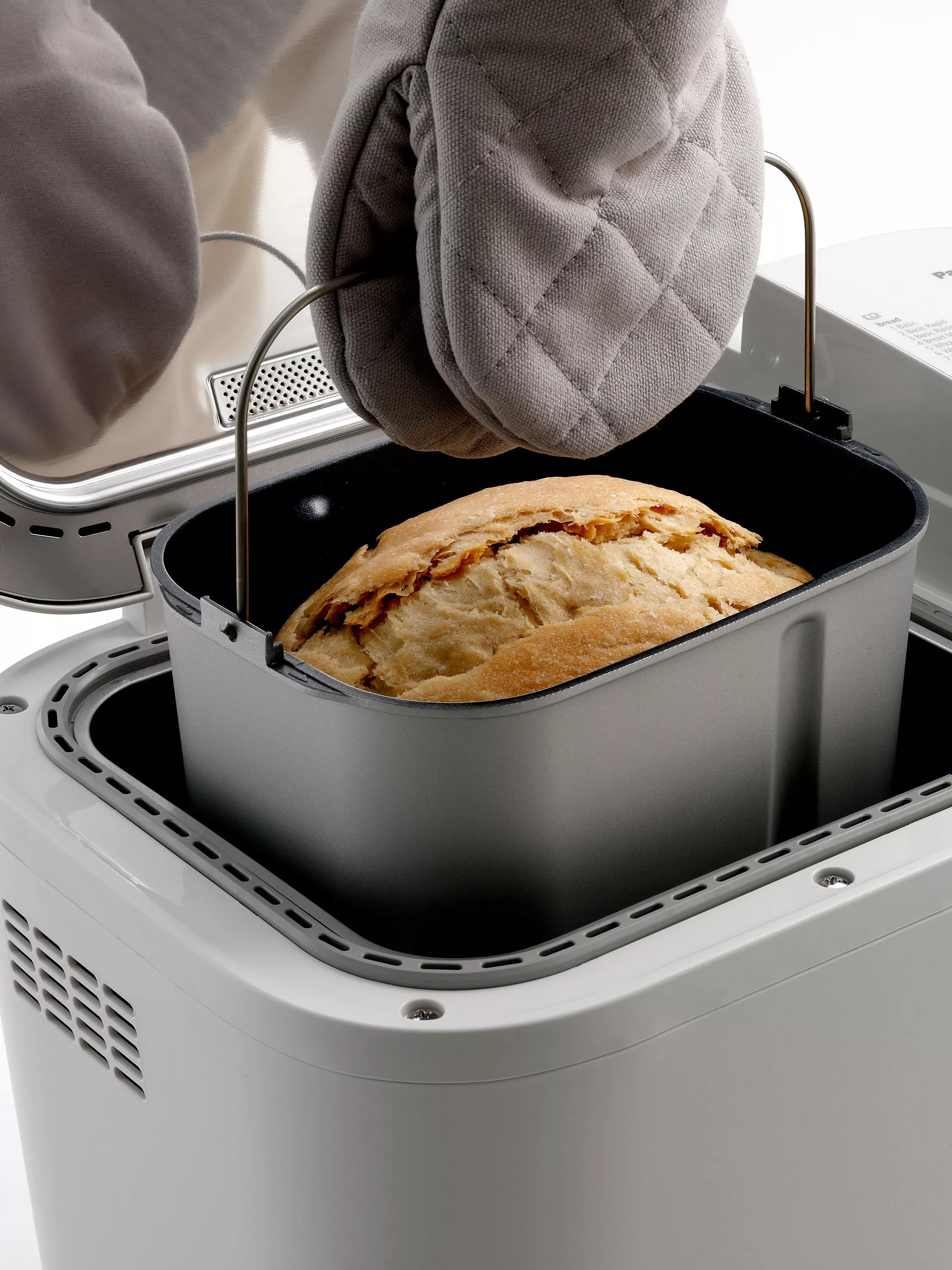 Freshly baked loaf in a bread maker tin, lifted with oven glove, homemade bread close-up in kitchen
