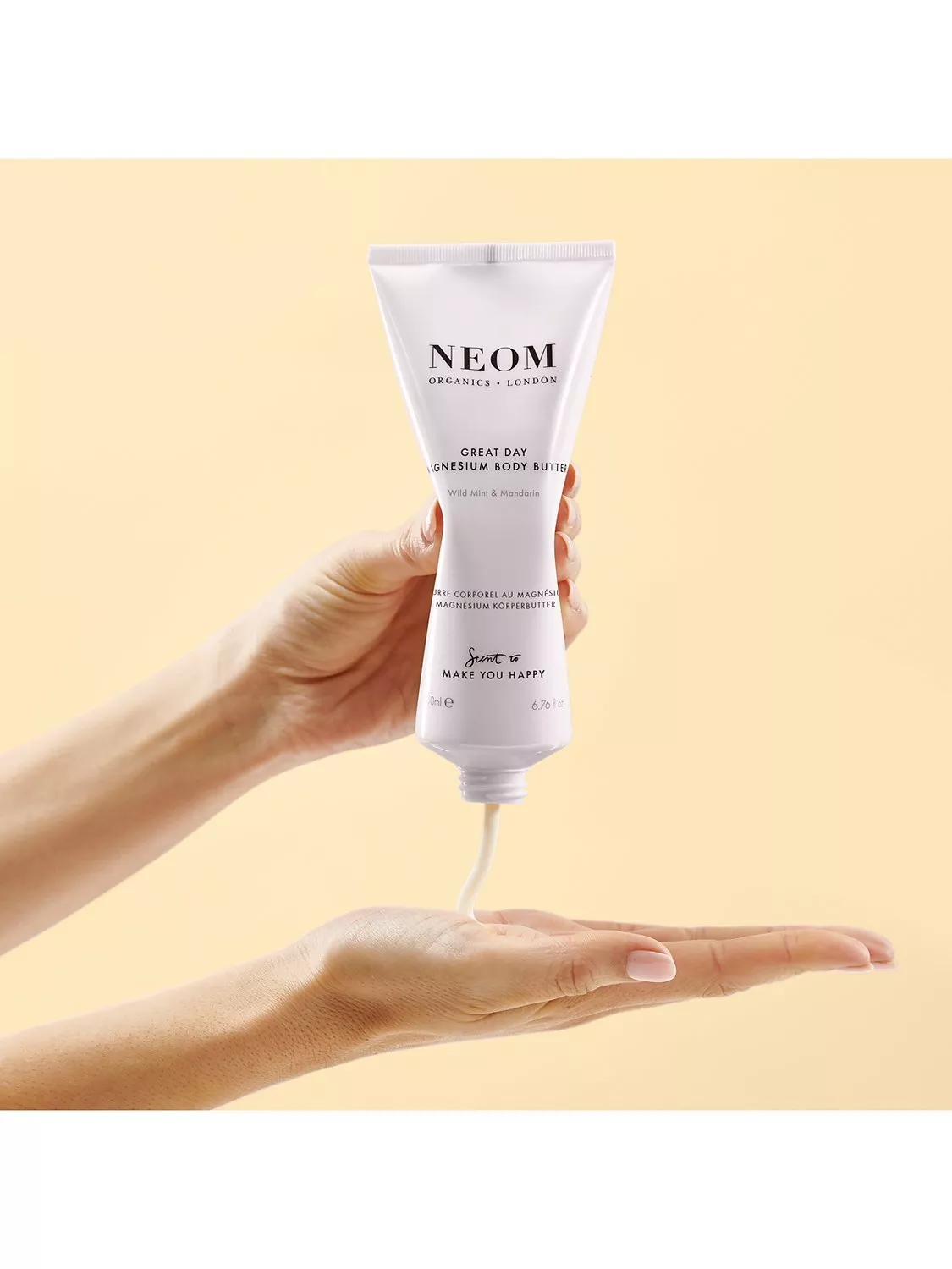 Hand holding NEOM Organics London Great Day magnesium body butter, dispensing lotion into palm on pastel background
