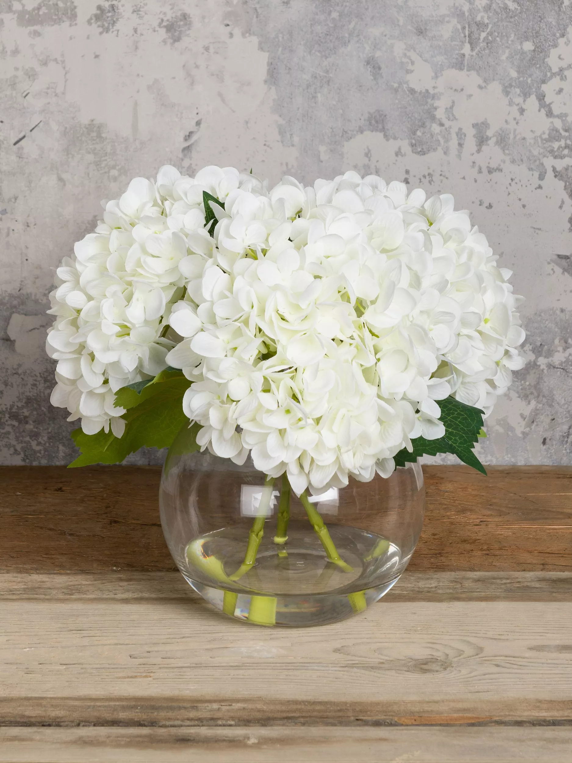 White hydrangea flower arrangement in a clear glass bowl vase on a rustic wooden table, grey textured wall backdrop