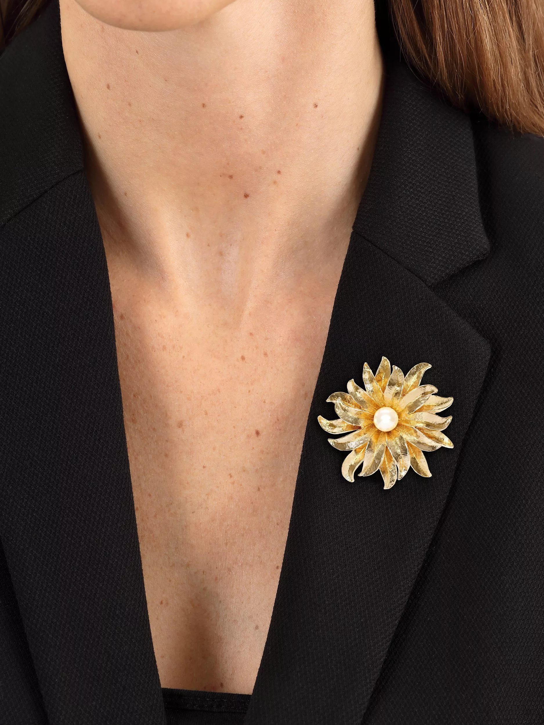 A gold floral brooch with a pearl centre adorns a black blazer, adding elegance to the neckline.