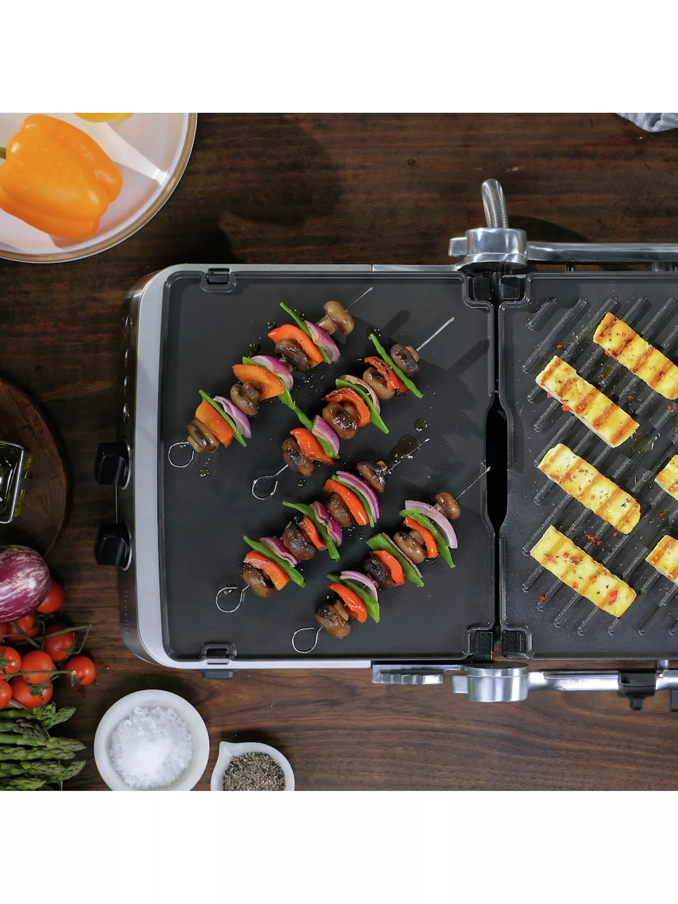 Overhead view of electric griddle cooking vegetable skewers and halloumi, with fresh veg and seasoning bowls.