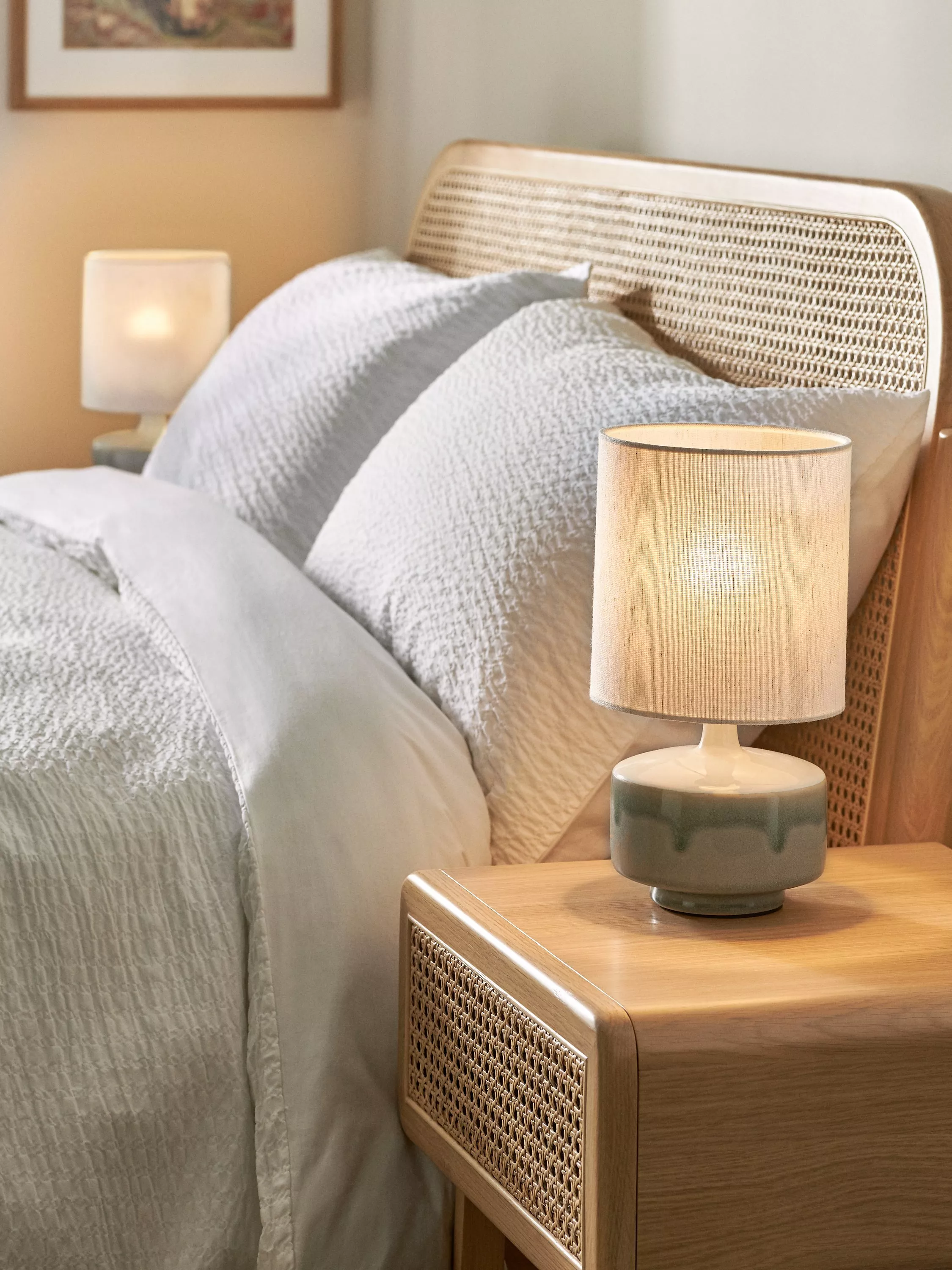 Cosy bedroom scene with rattan headboard, wooden bedside table and ceramic lamp beside crisp white bedding