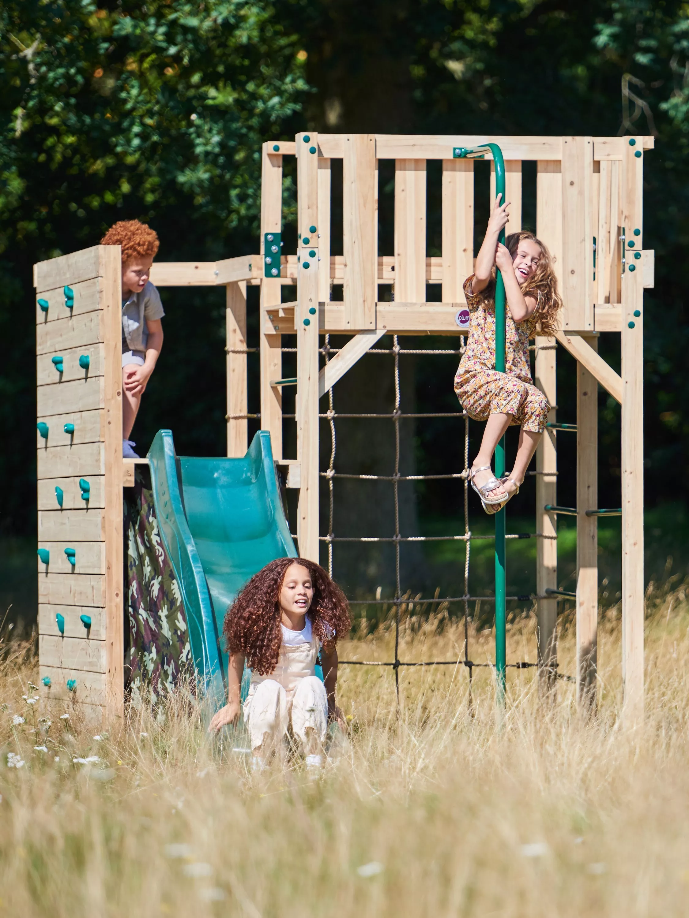 Children enjoying a wooden playground with a slide and climbing structures in a grassy park setting.