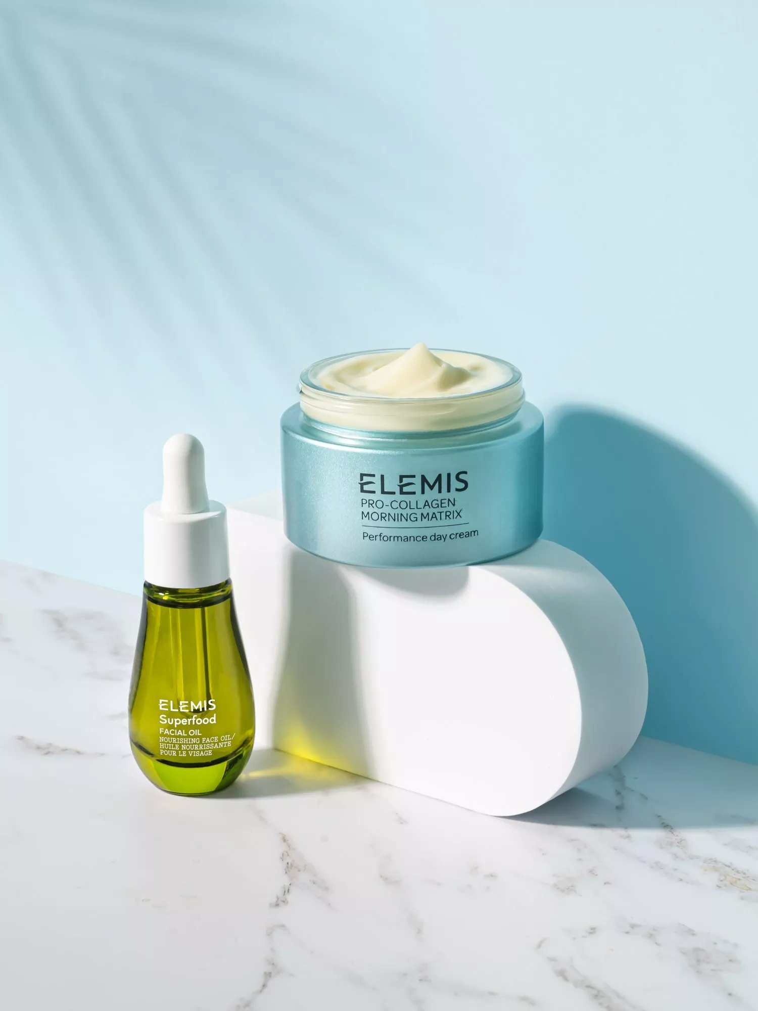 Skincare products on a marble surface, featuring Elemis Pro-Collagen cream and Superfood facial oil in bright light.