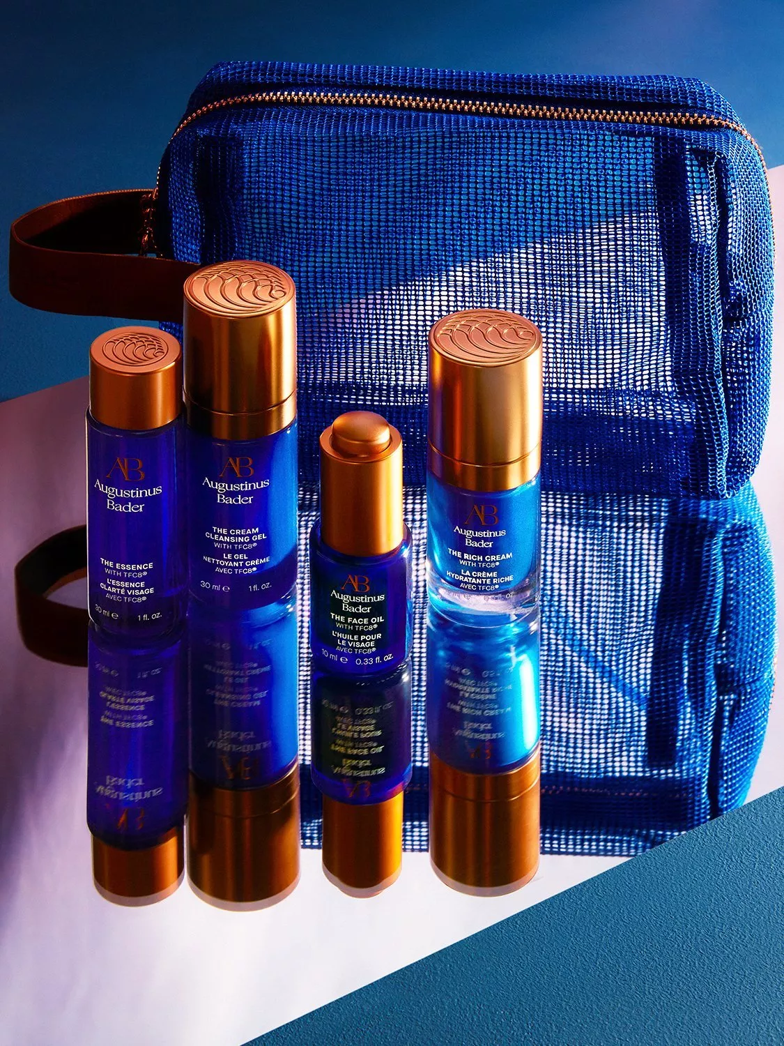 Augustinus Bader blue skincare bottles with gold caps beside a blue mesh cosmetic wash bag on a surface