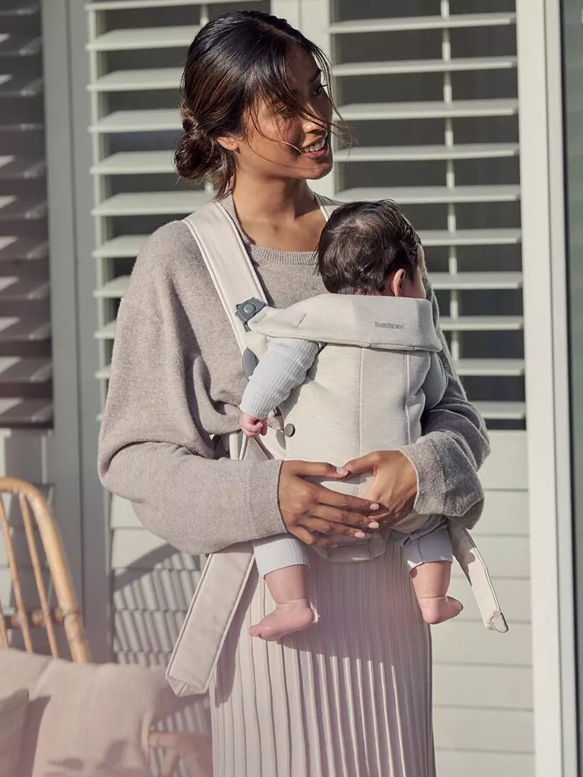 Smiling woman carrying baby in beige front baby carrier outdoors by window shutters in soft sunlight