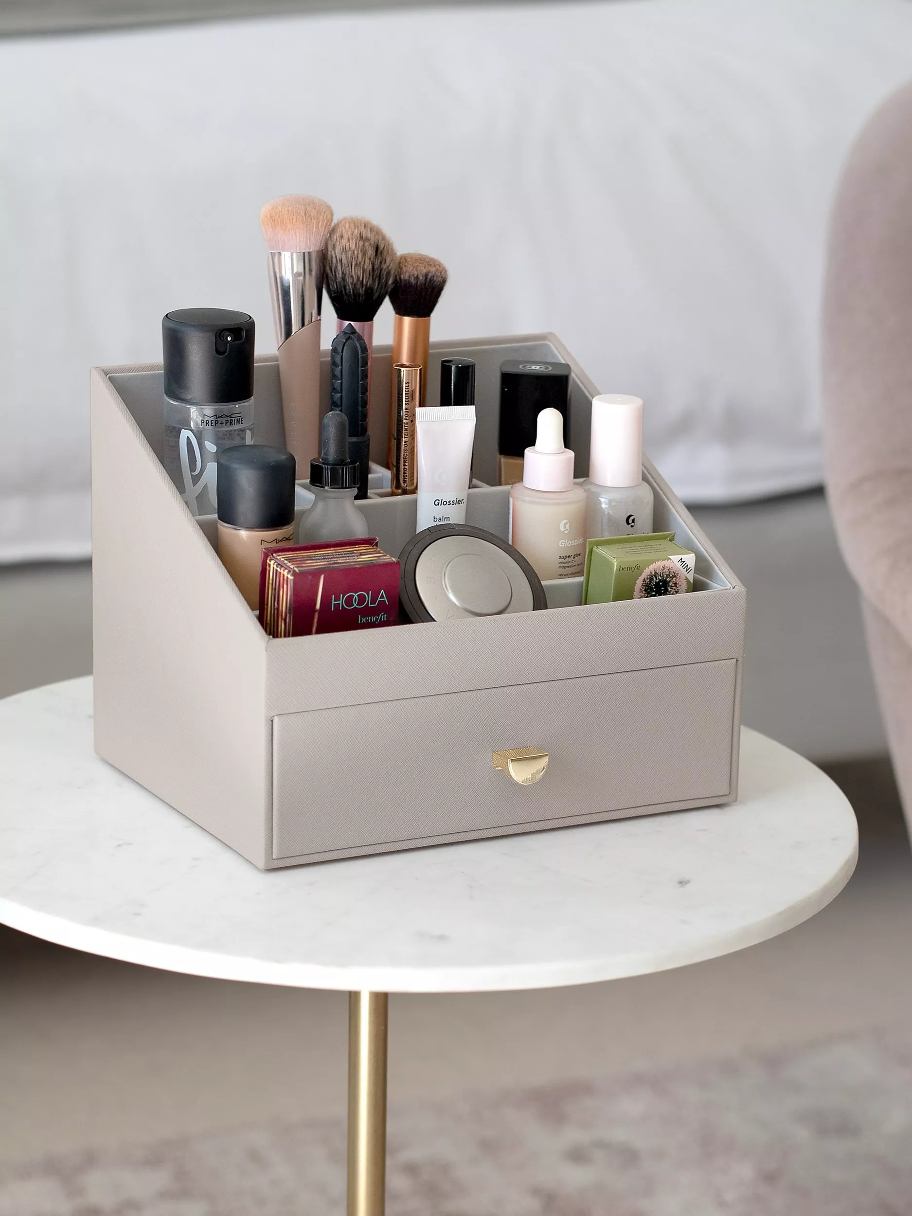 Grey makeup organiser box with drawer, holding brushes and cosmetics, on a round marble side table