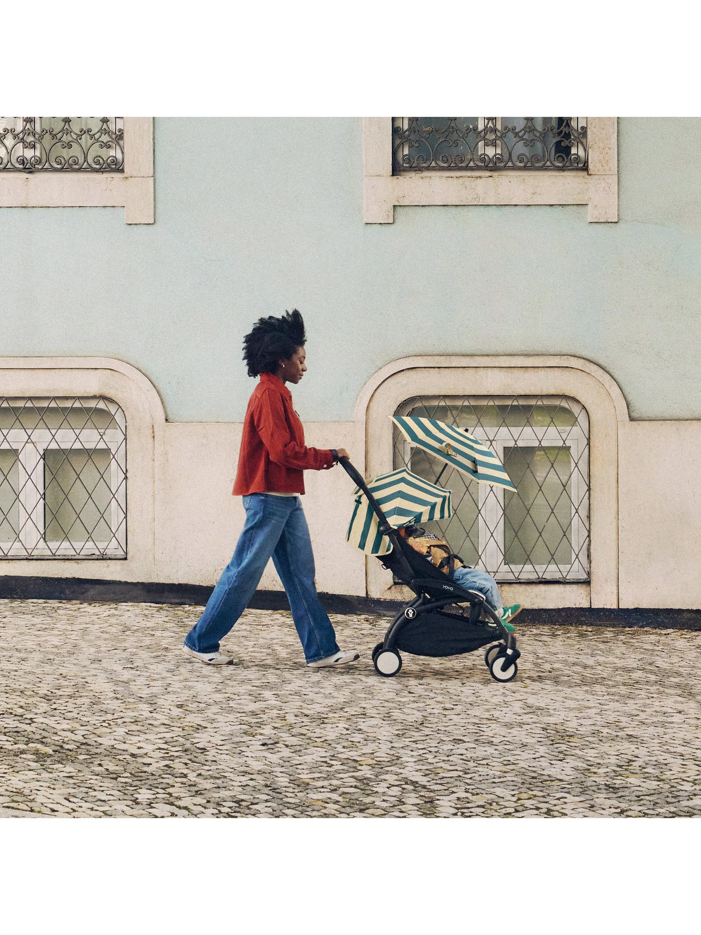 Woman pushing baby stroller with striped sun canopy on cobbled street by pastel building facade outdoors