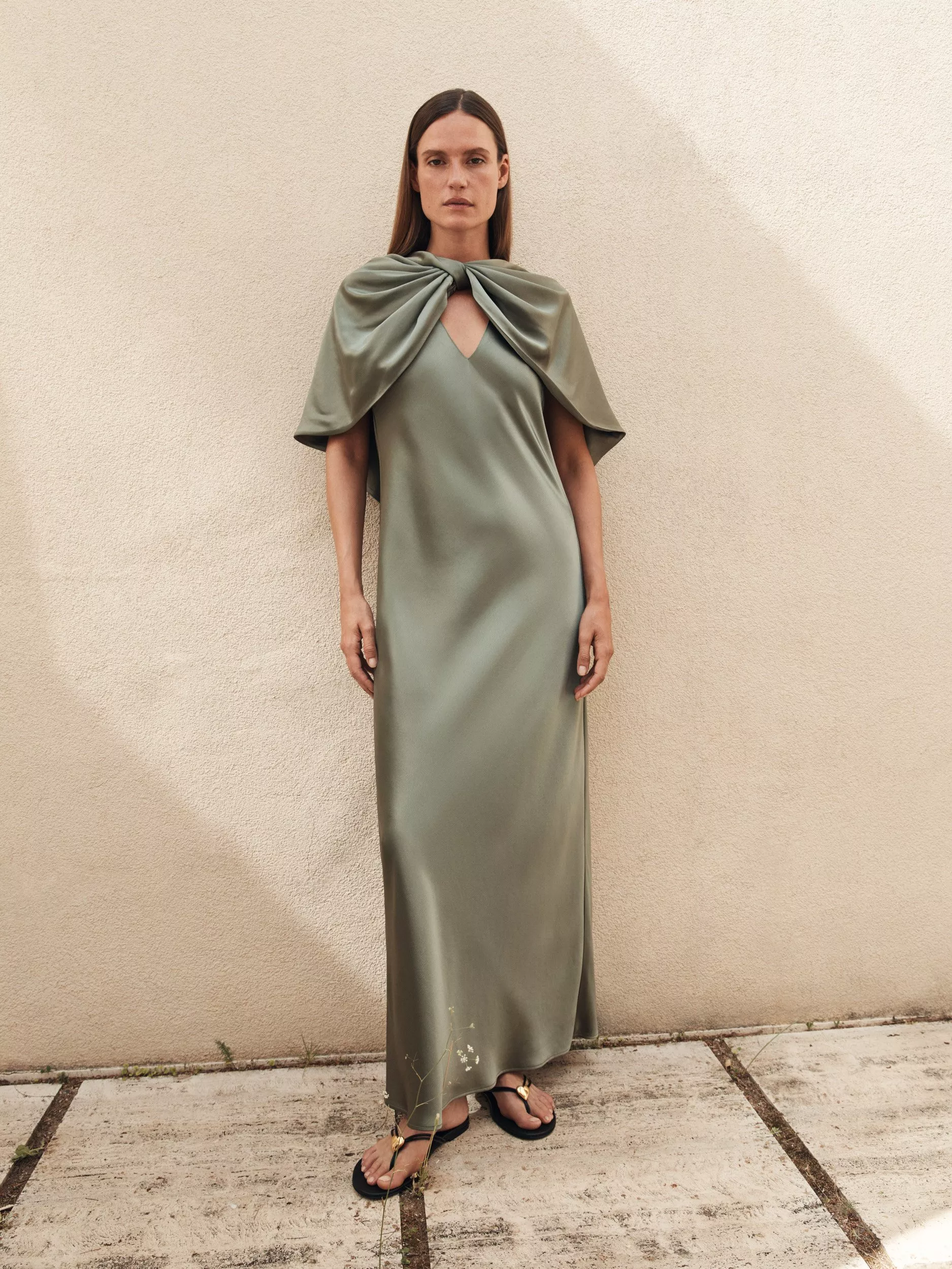 Woman wearing a sage green satin maxi dress with draped cape detail, standing against a beige wall outdoors.