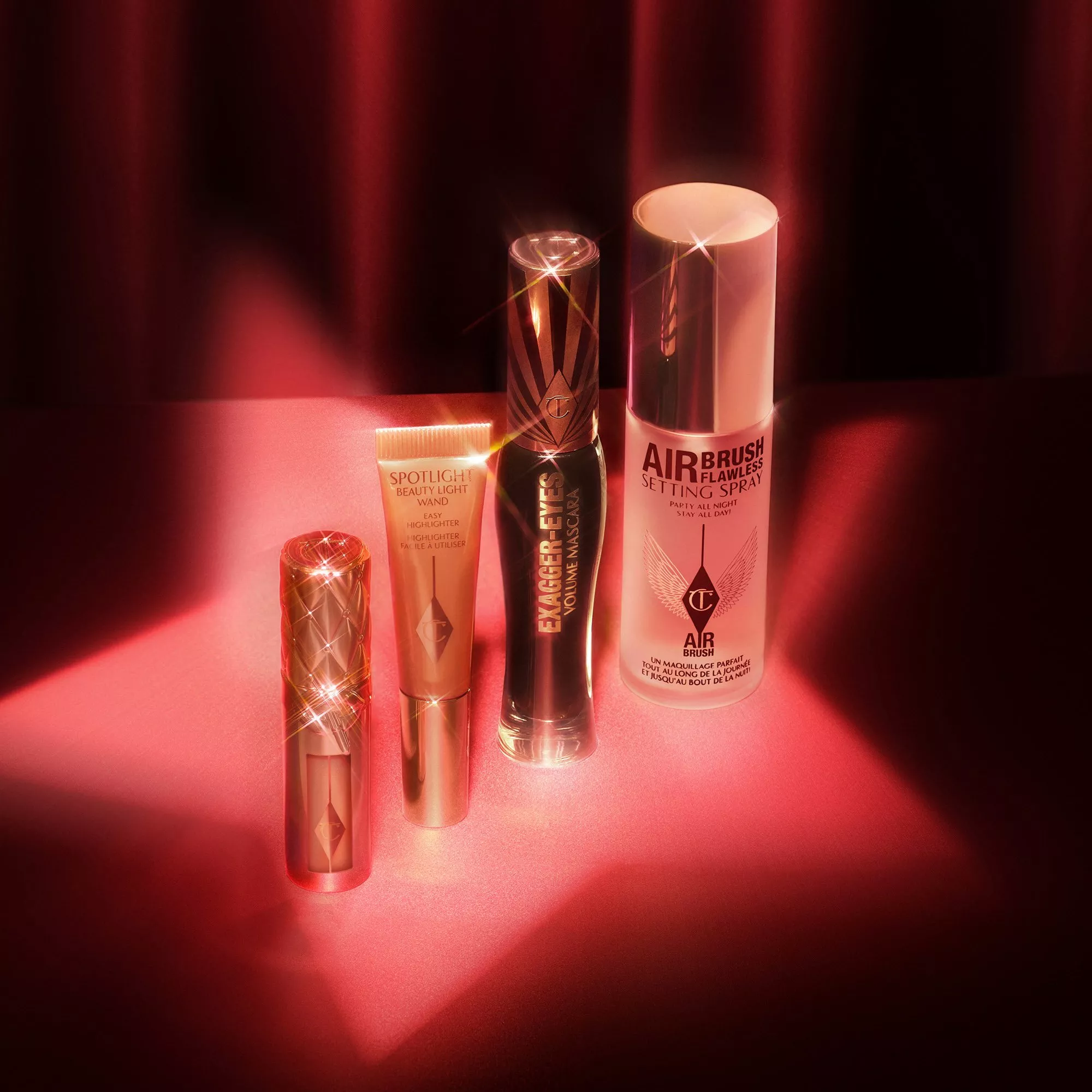 Charlotte Tilbury makeup set with setting spray, mascara, highlighter and lipstick on red backdrop, studio lighting