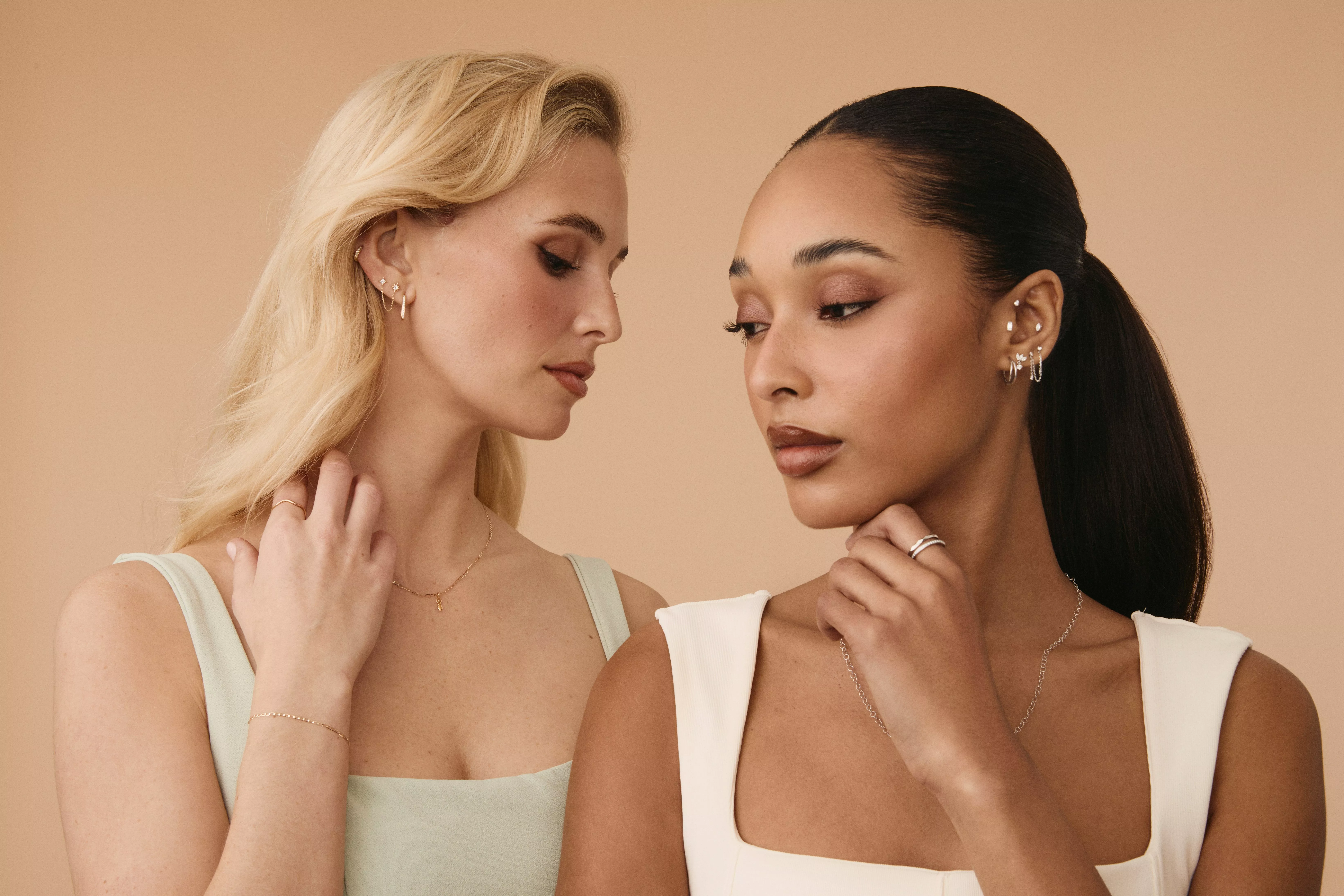 Two women modelling delicate gold earrings and rings, beauty portrait with natural make-up on beige background
