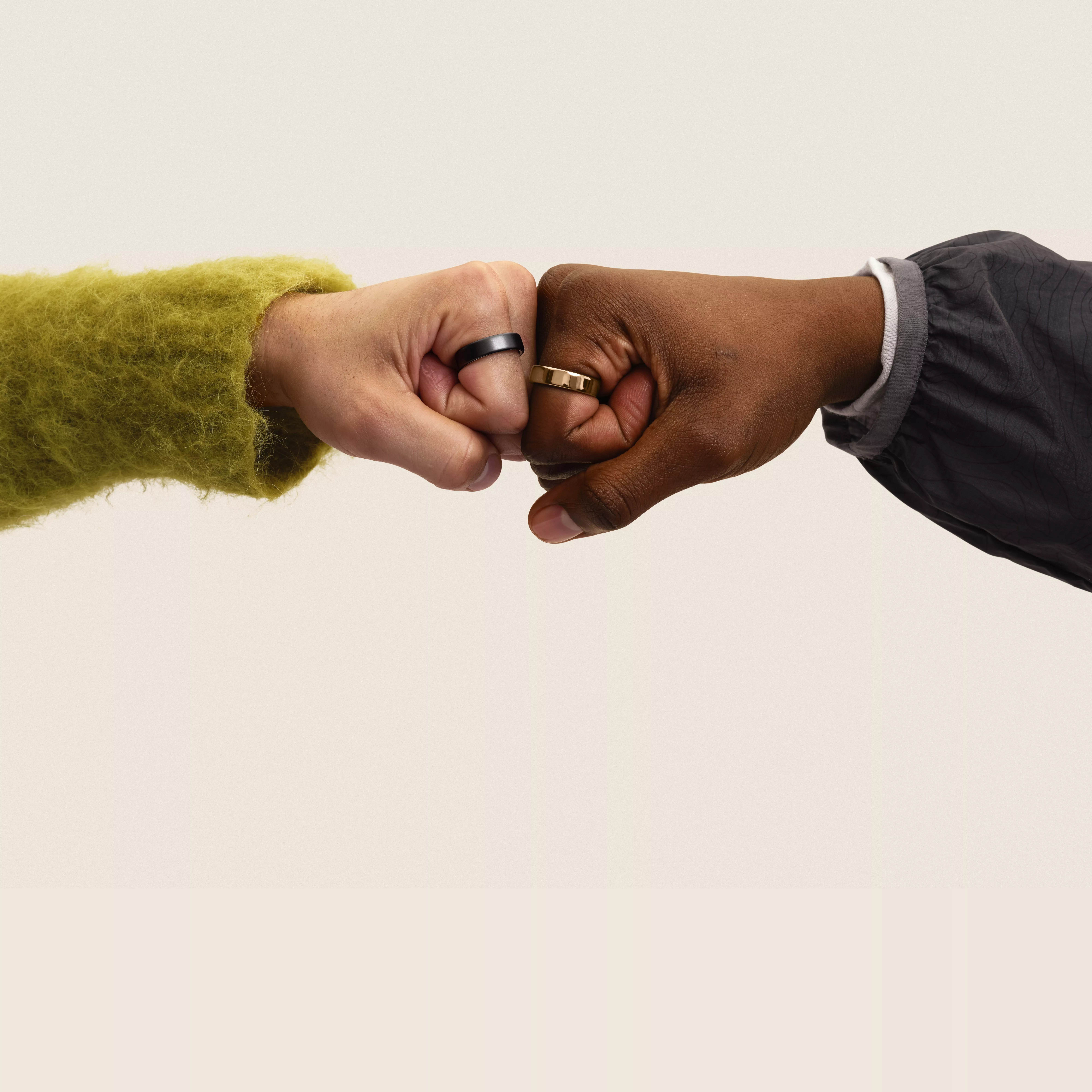 Two hands side-by-side using their index and middle fingers to "walk" like legs. One wears a gold Oura ring and dark jacket sleeve and the other wears a black Oura ring and a chartreuse jumper sleeve.
