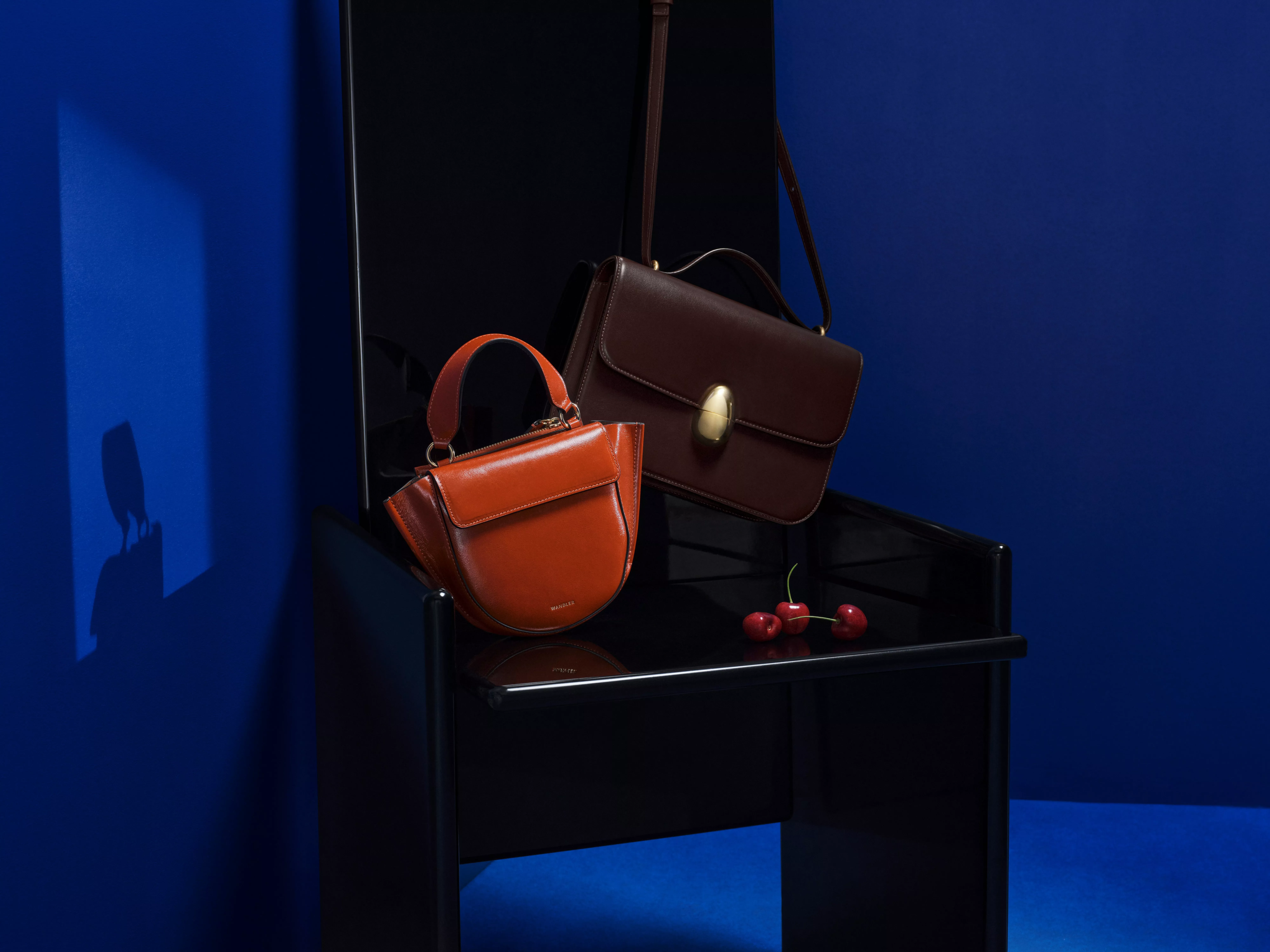 Luxury leather handbags on glossy black chair, brown crossbody and tan top-handle bag against blue backdrop