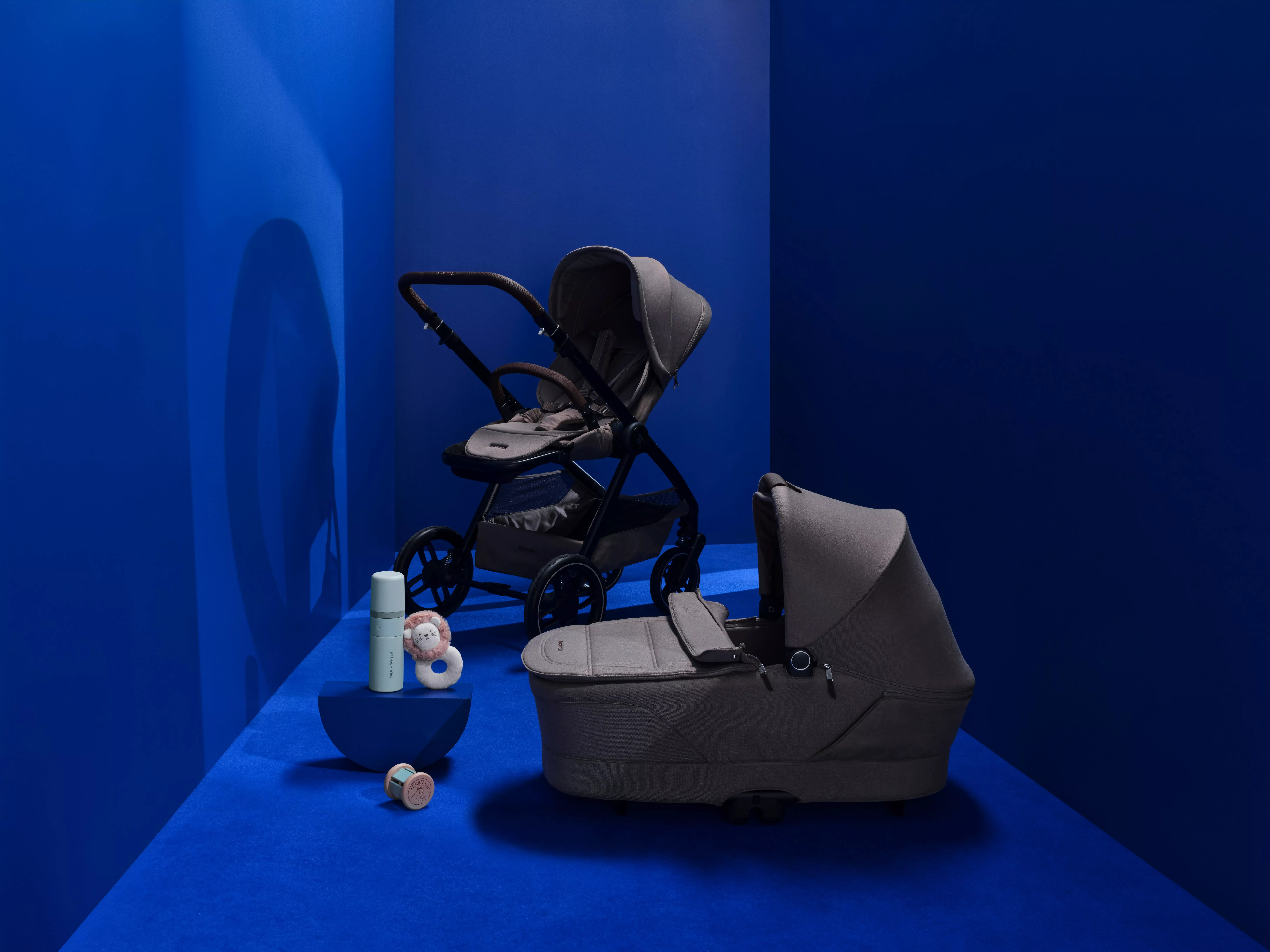 Grey baby pushchair and carrycot pram set in a blue studio, with toy and bottle, modern travel system setup