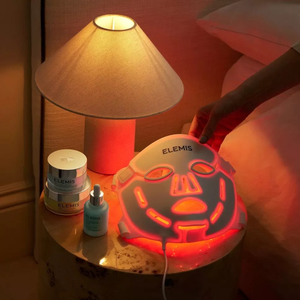 ELEMIS LED face mask glowing red on bedside table with skincare jars and lamp in cosy bedroom setting