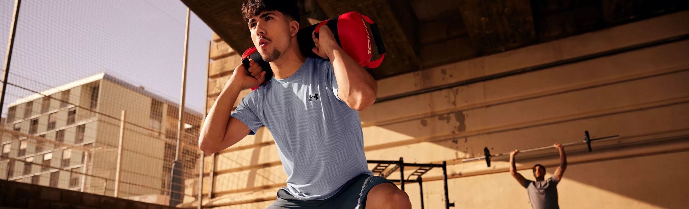 man doing outdoor fitness training with a sandbag on shoulders under an urban bridge in sunlight