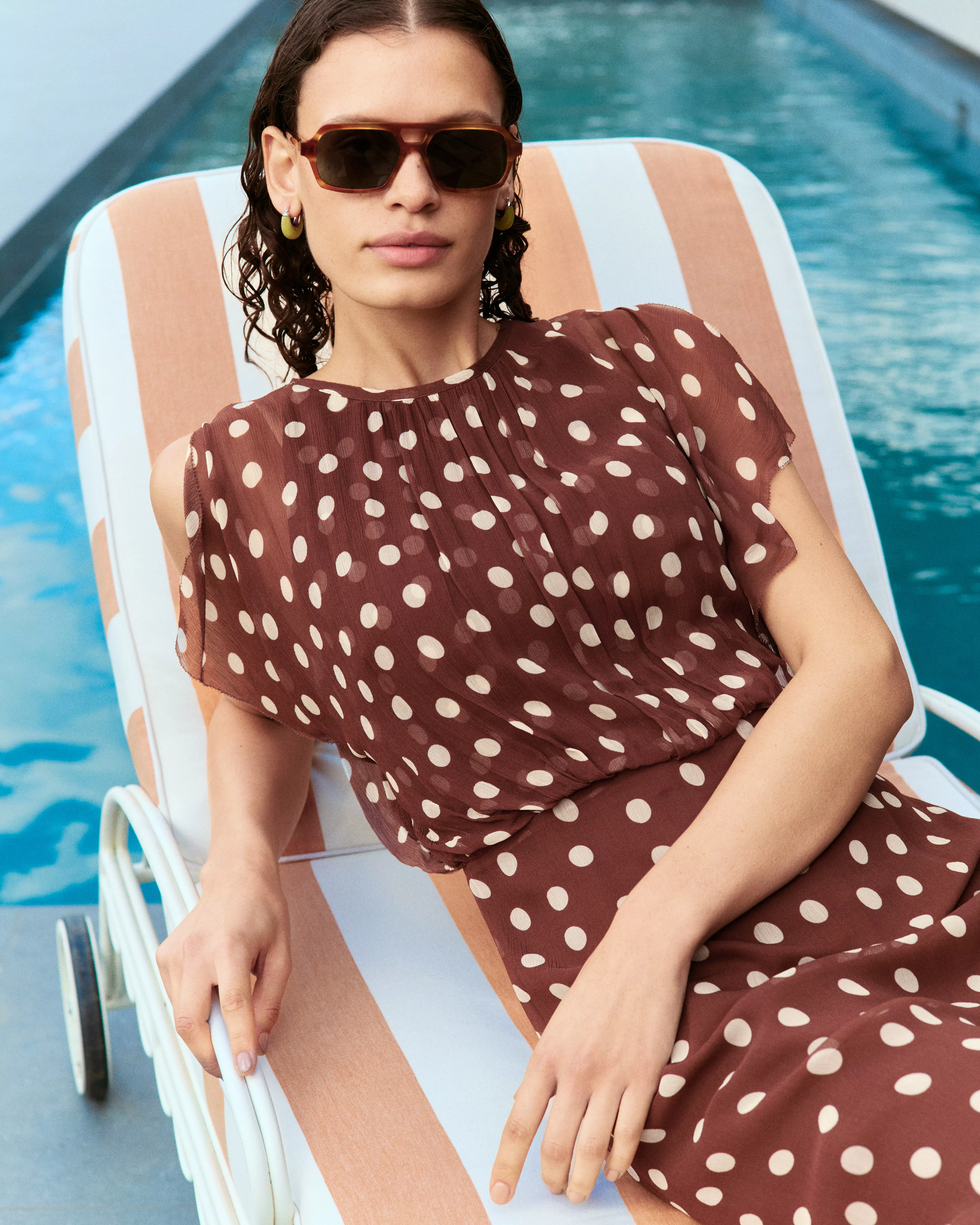 A person wearing sunglasses lounges by a pool on a striped chair. They sport a brown polka dot dress.