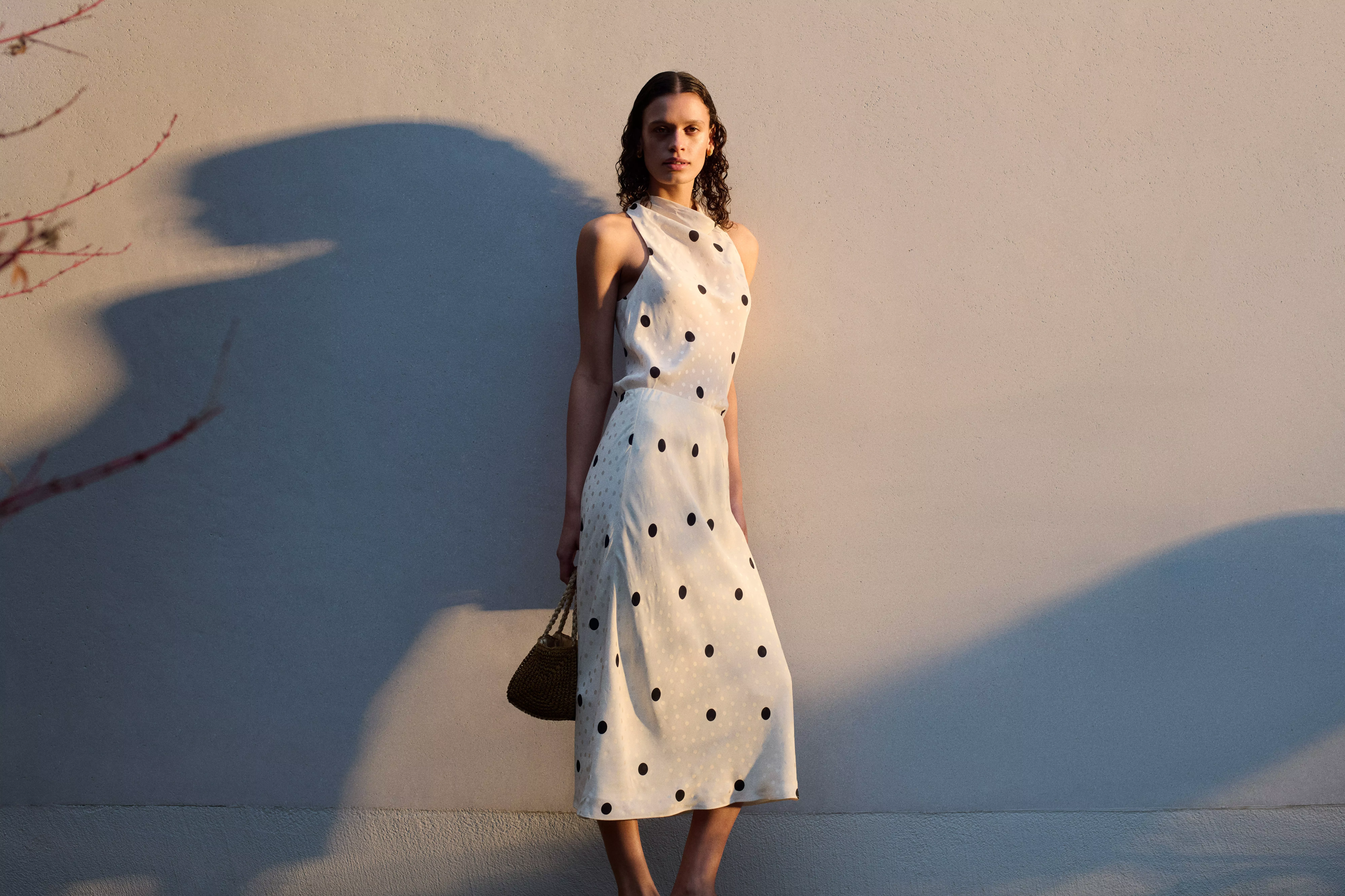 A woman in a sleeveless, polka-dot dress stands against a plain wall, holding a small handbag in warm lighting.