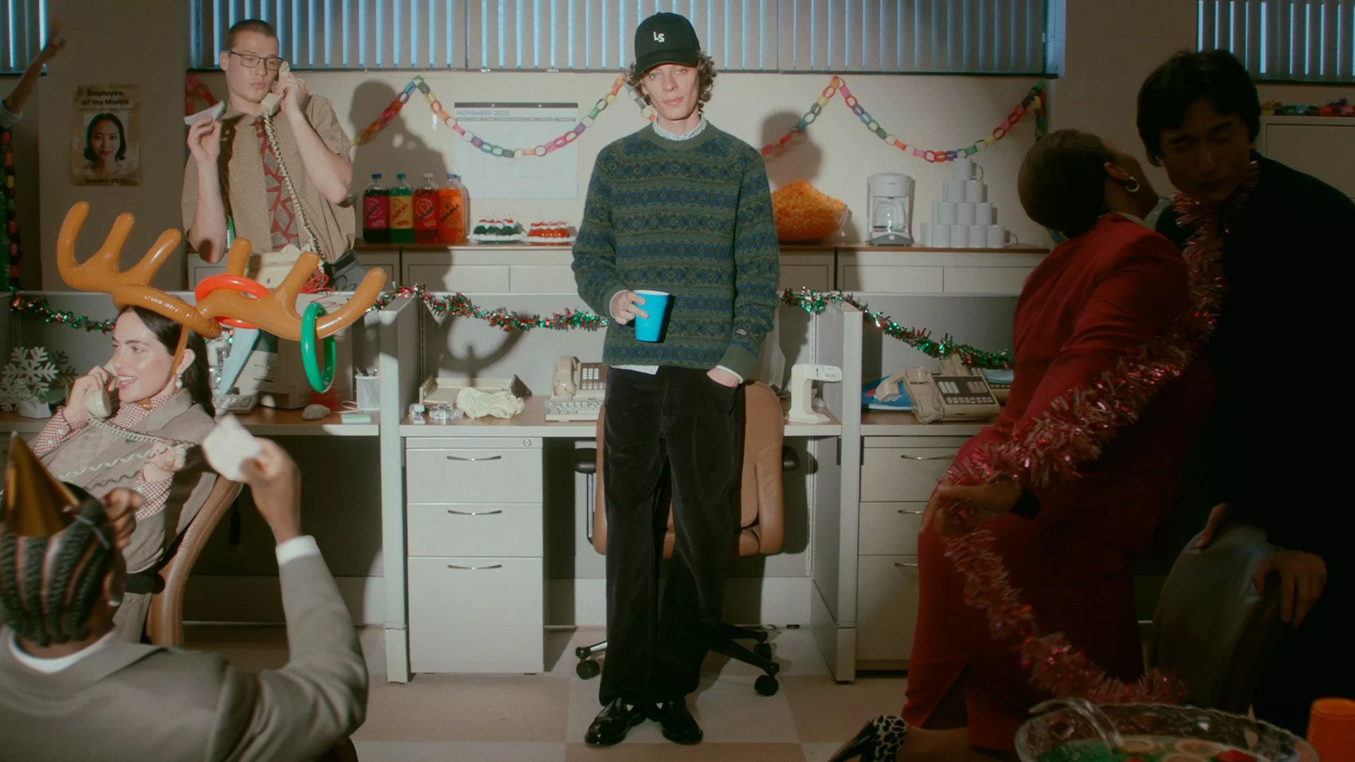 An awkward holiday office party scene. A young man, wearing a baseball cap and a patterned sweater, holds a blue cup and looks directly at the camera, surrounded by casually dressed co-workers celebrating with tinsel and festive decorations.