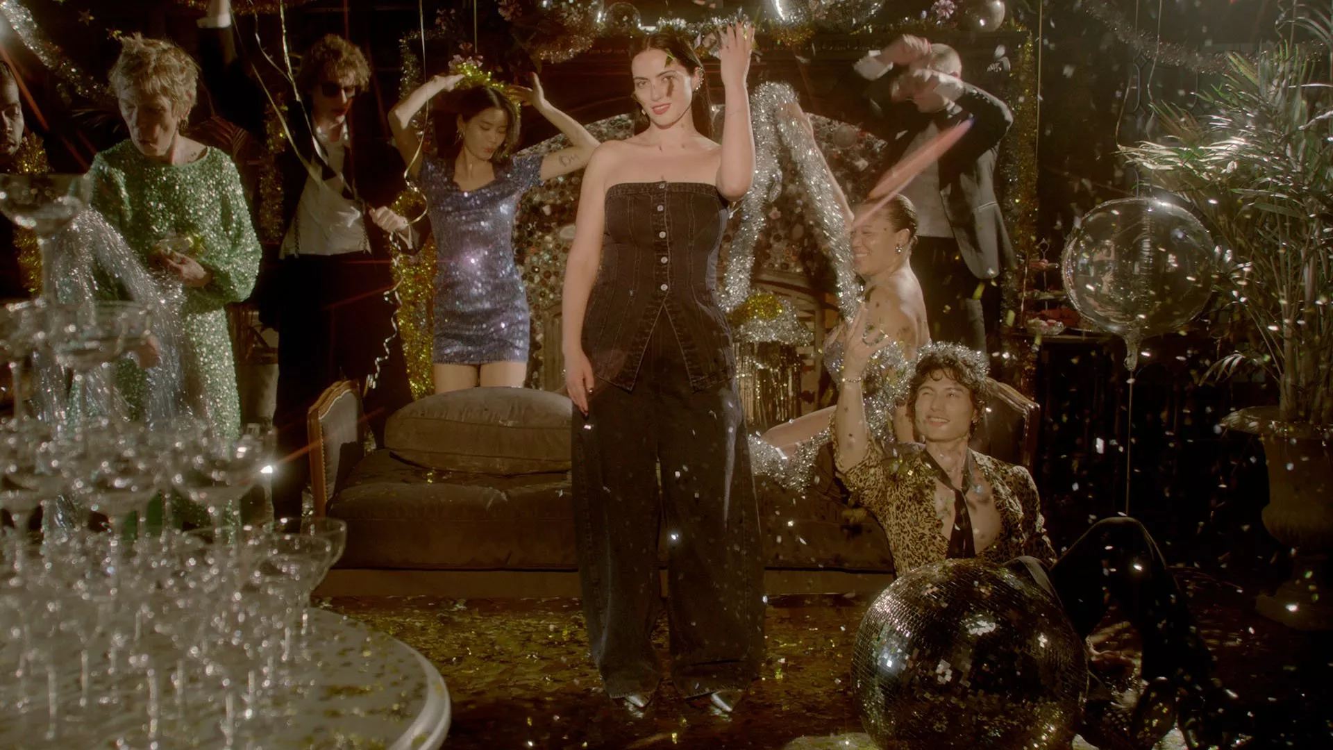 A dazzling holiday party scene filled with glitter and confetti. People are dressed in festive, shiny attire, including a woman in the center wearing an all-denim strapless jumpsuit, and another person sitting on the floor next to a large disco ball.