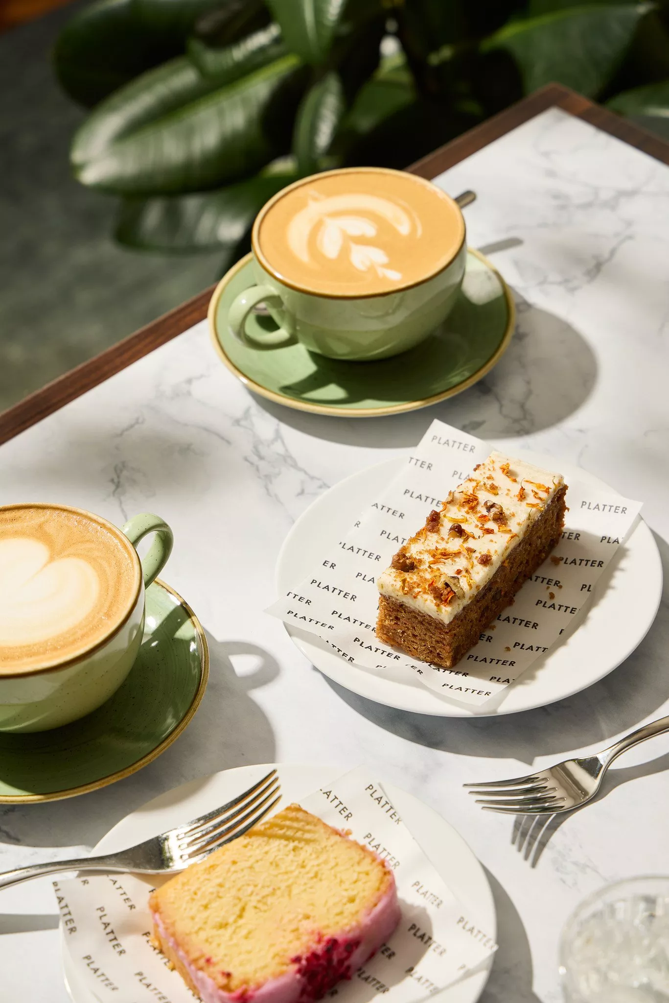 Two lattes in green cups with latte art, served with cake slices on a marble café table in sunlight