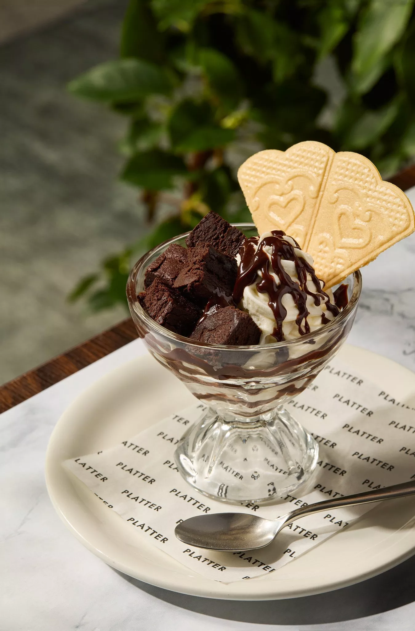 Chocolate brownie sundae with vanilla ice cream, chocolate sauce and heart biscuit in a glass dessert dish