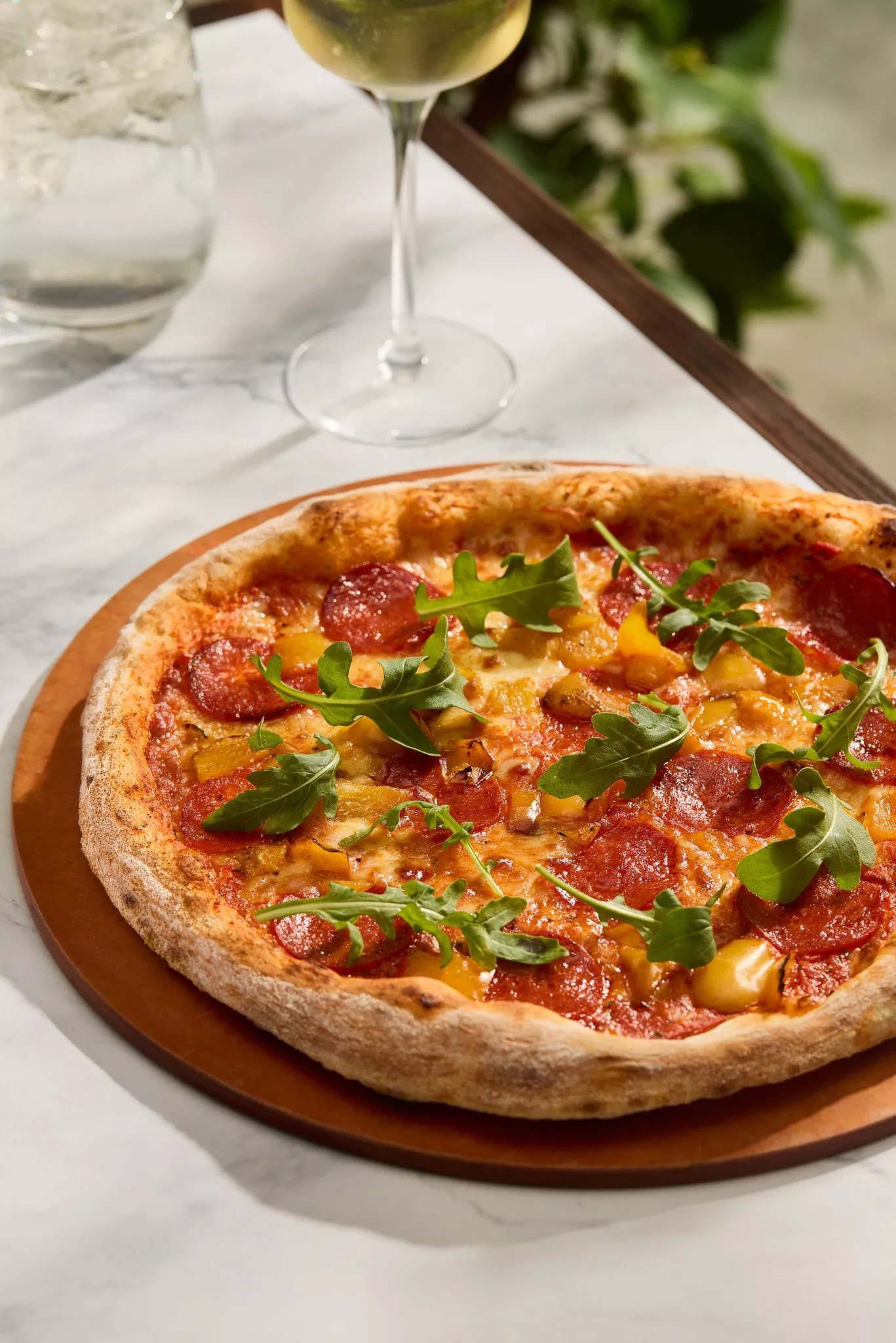 Pepperoni pizza with pineapple and rocket on a wooden board, served with a glass of white wine on marble table